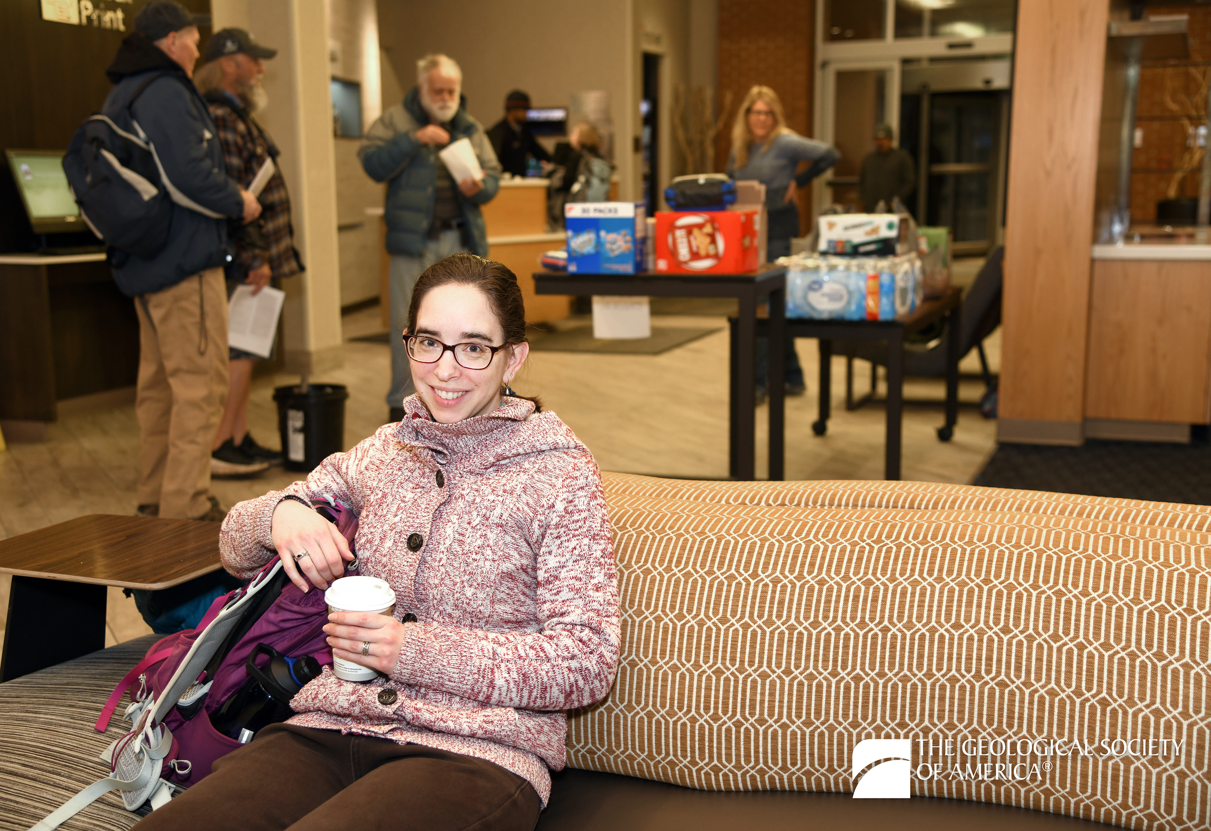 A woman in a cozy sweater sits on a patterned couch, smiling and holding a coffee. Behind her, people gather near a table with snacks and drinks in a welcoming indoor setting.