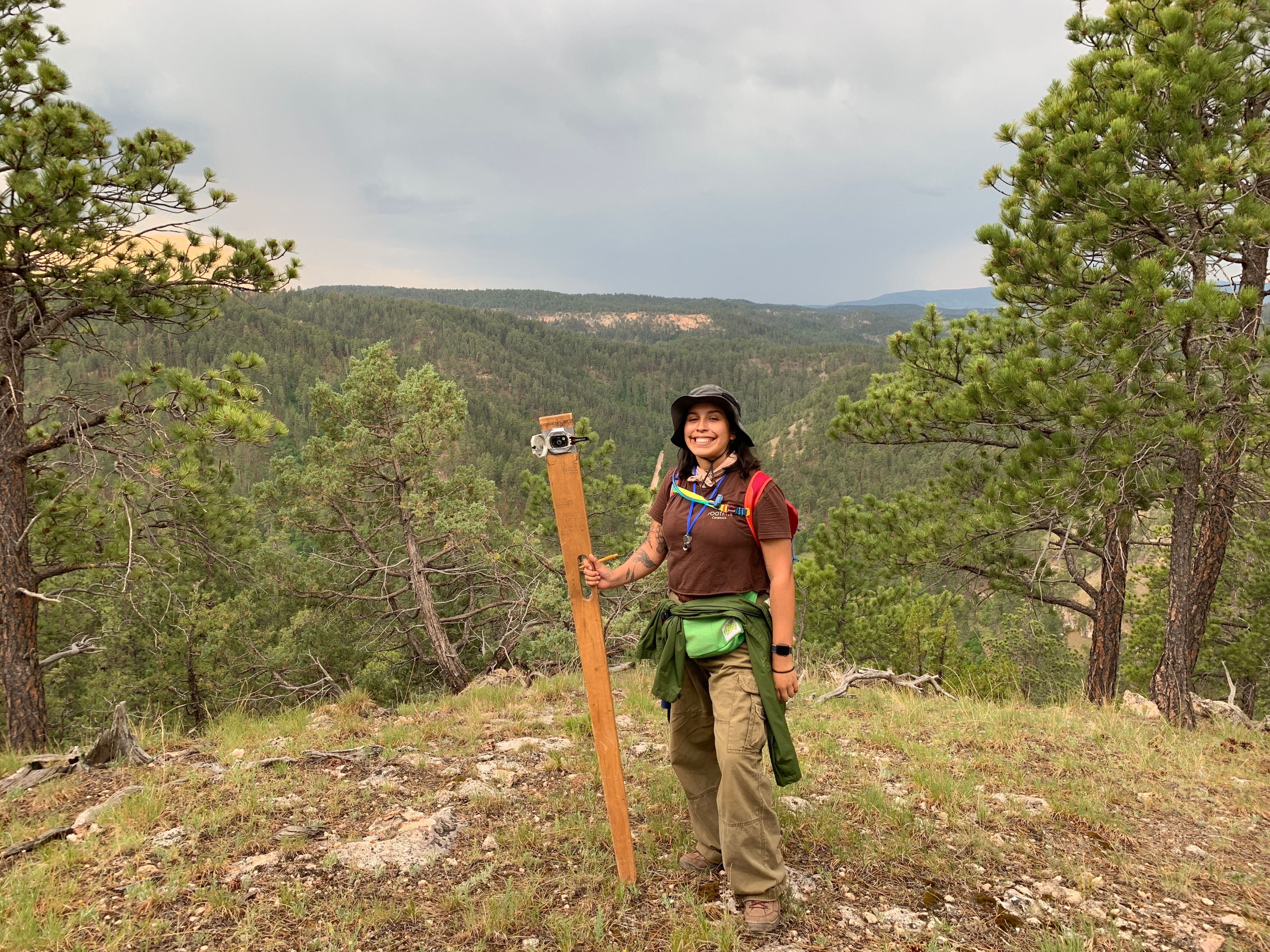 A smiling woman in hiking gear and a sun hat stands on a forested ridge, holding a wooden Jacob’s staff used for geological measuring. A vast, evergreen-covered valley stretches out behind her under a cloudy sky.