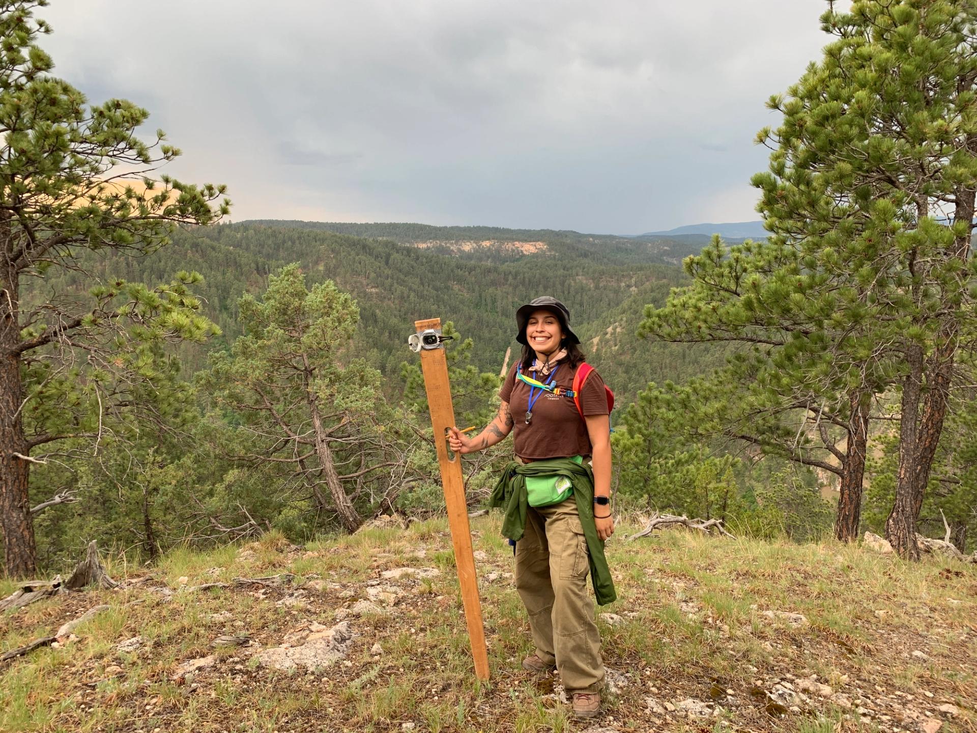 A smiling woman in hiking gear and a sun hat stands on a forested ridge, holding a wooden Jacob’s staff used for geological measuring. A vast, evergreen-covered valley stretches out behind her under a cloudy sky.
