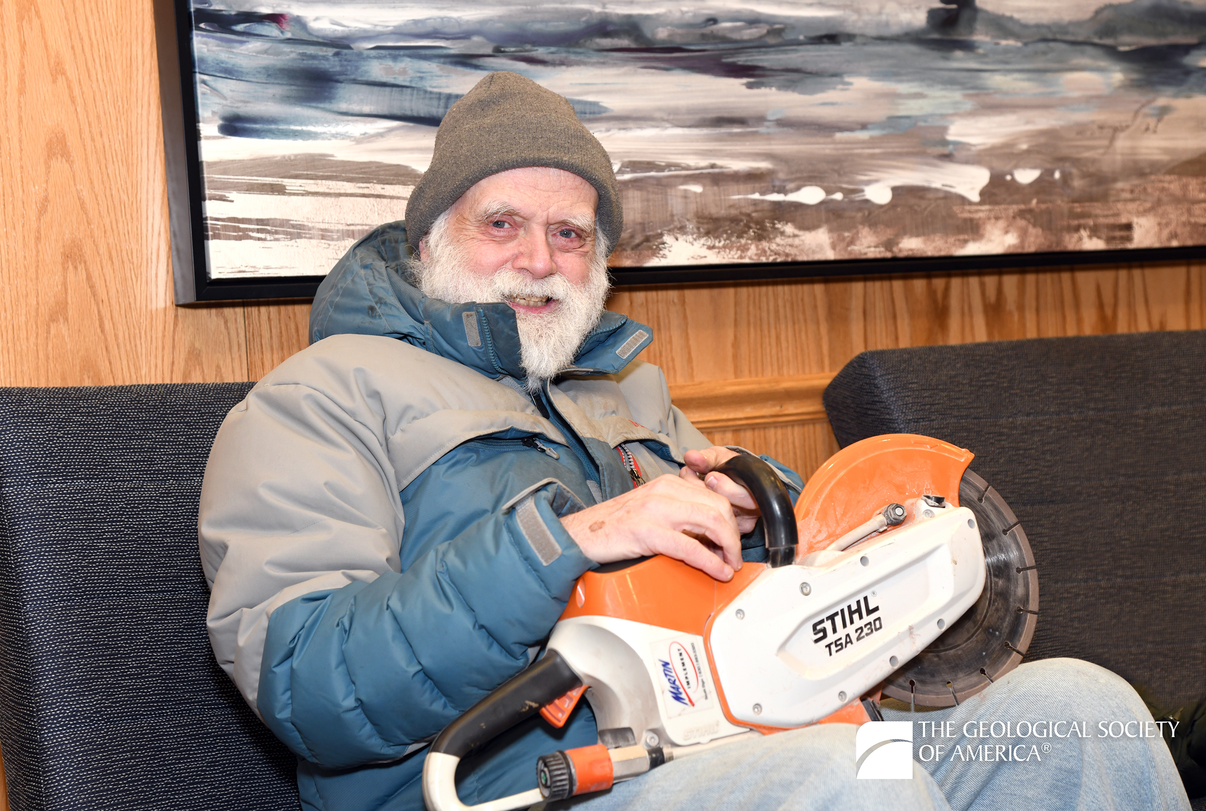 An elderly man in a winter jacket and beanie smiles while holding an orange STIHL chainsaw. The background features abstract art and wood paneling. The atmosphere is cozy and relaxed.