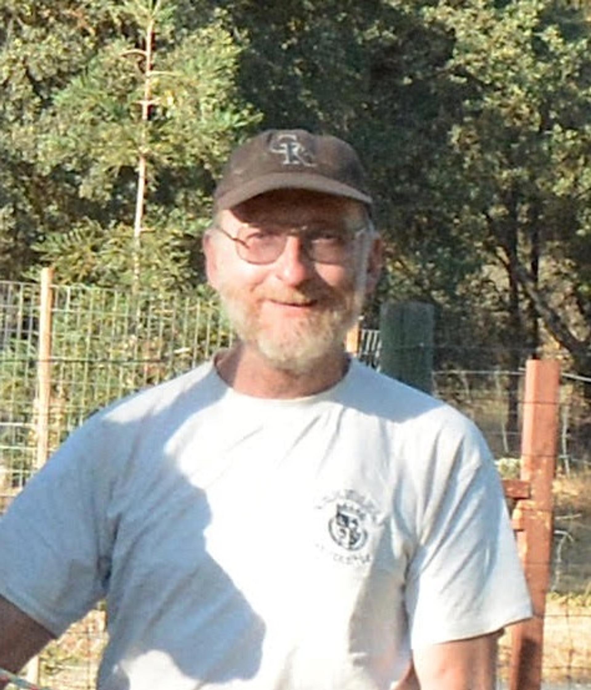 Craig H. Jones in a white t-shirt and dark baseball cap with wire fencing and green trees in the background.