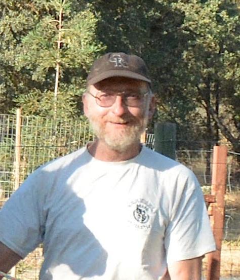 Craig H. Jones in a white t-shirt and dark baseball cap with wire fencing and green trees in the background.