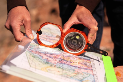 A person holds an orange Brunton orange tool against a map. They are outdoors.