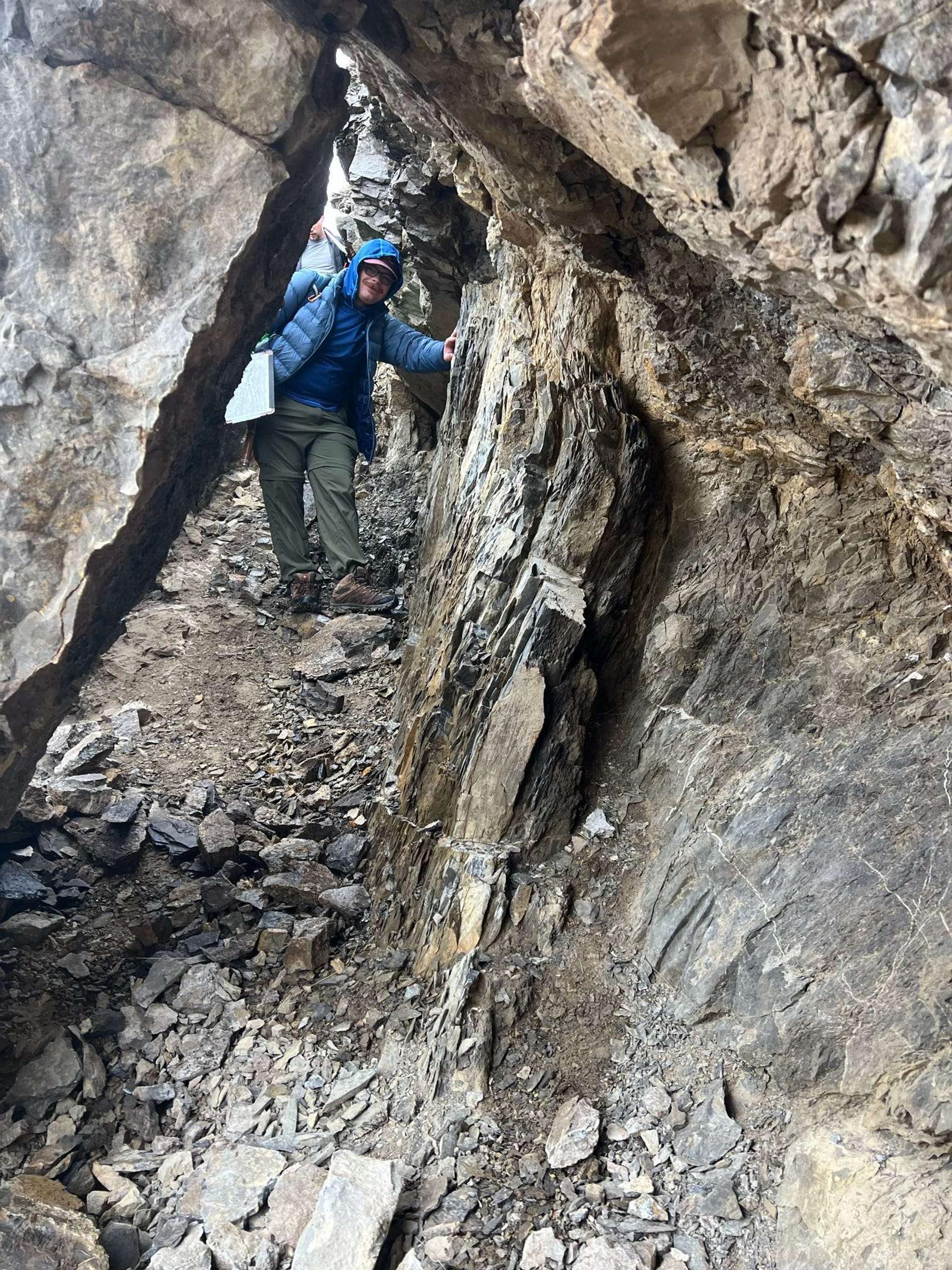 A man in a blue jacket and hiking pants walks through a narrow rock tunnel.