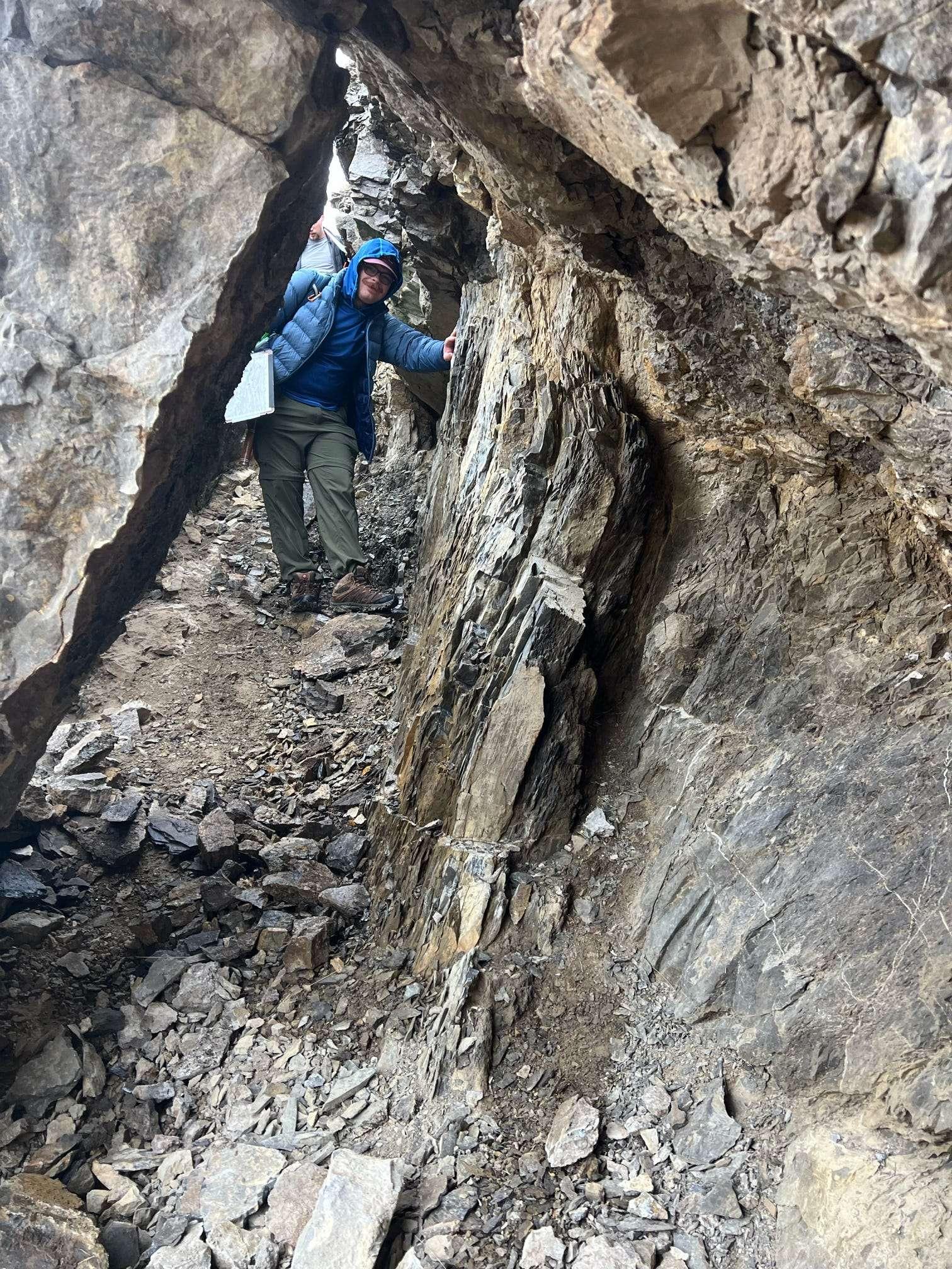 A man in a blue jacket and hiking pants walks through a narrow rock tunnel.