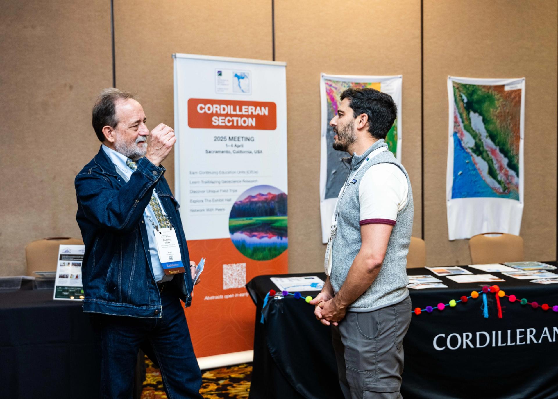 Two men engage in conversation at a conference booth. Behind them, a "Cordilleran Section" banner and maps are displayed, creating a professional ambiance.