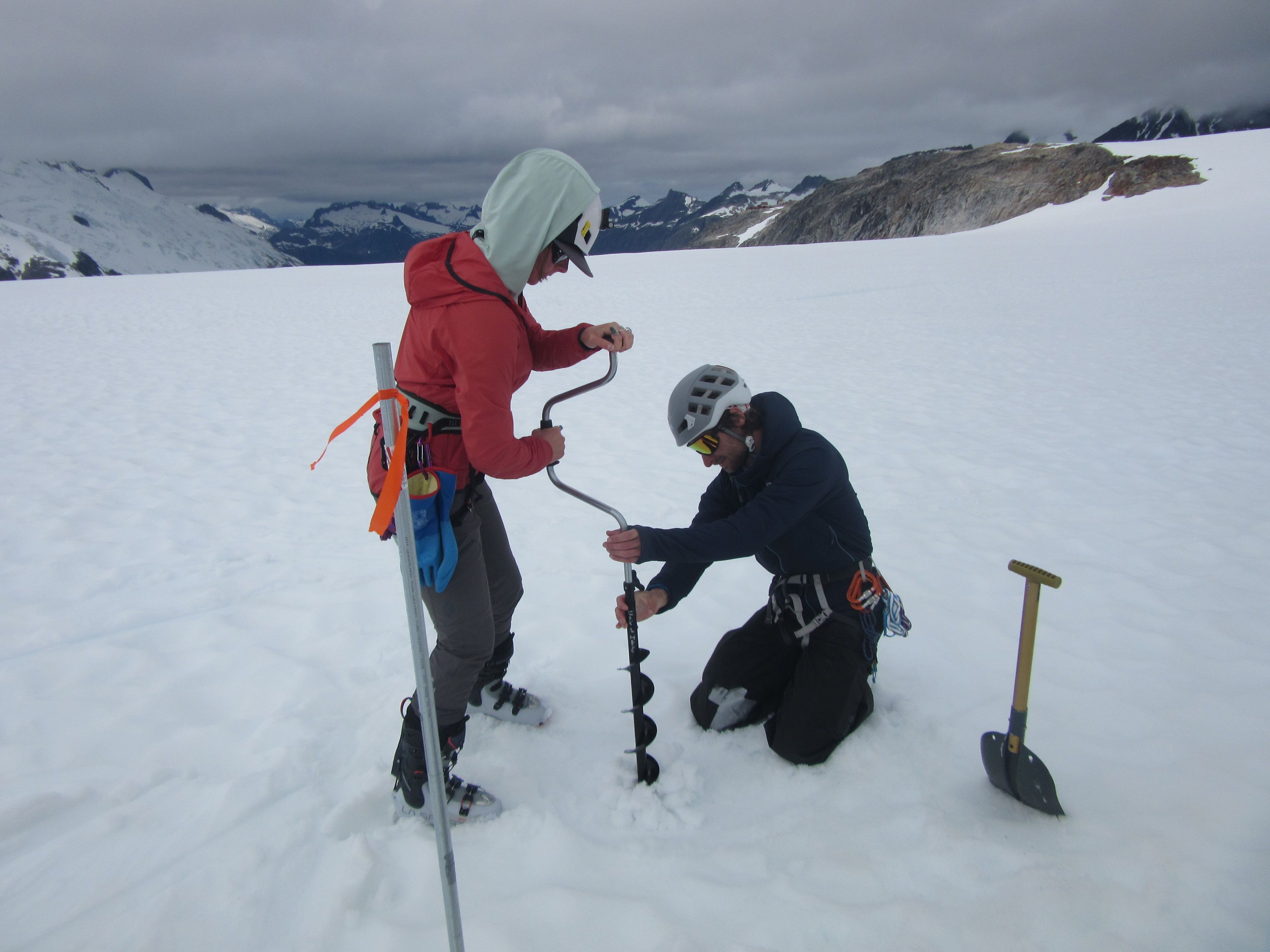 This is a pair of researchers on a glacier or ice field, likely conducting mass balance monitoring or firn density studies. They are using a manual ice auger to drill into the surface.
