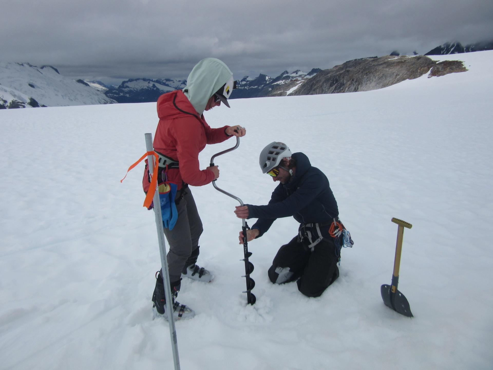 This is a pair of researchers on a glacier or ice field, likely conducting mass balance monitoring or firn density studies. They are using a manual ice auger to drill into the surface.