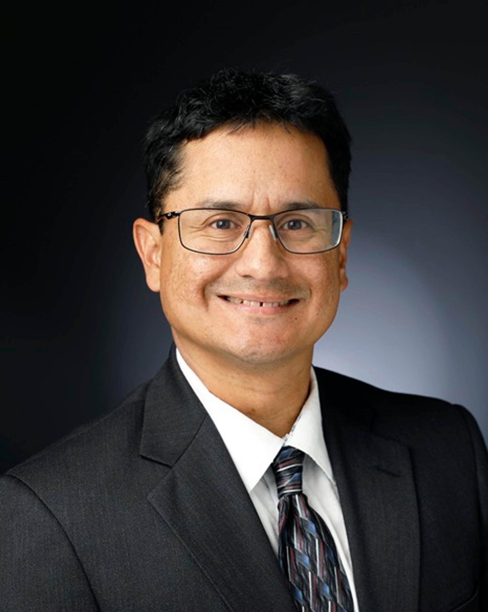 Jose M. Hurtado, Jr. in a dark suit jacket, tie, white shirt, and glasses against a dark background.