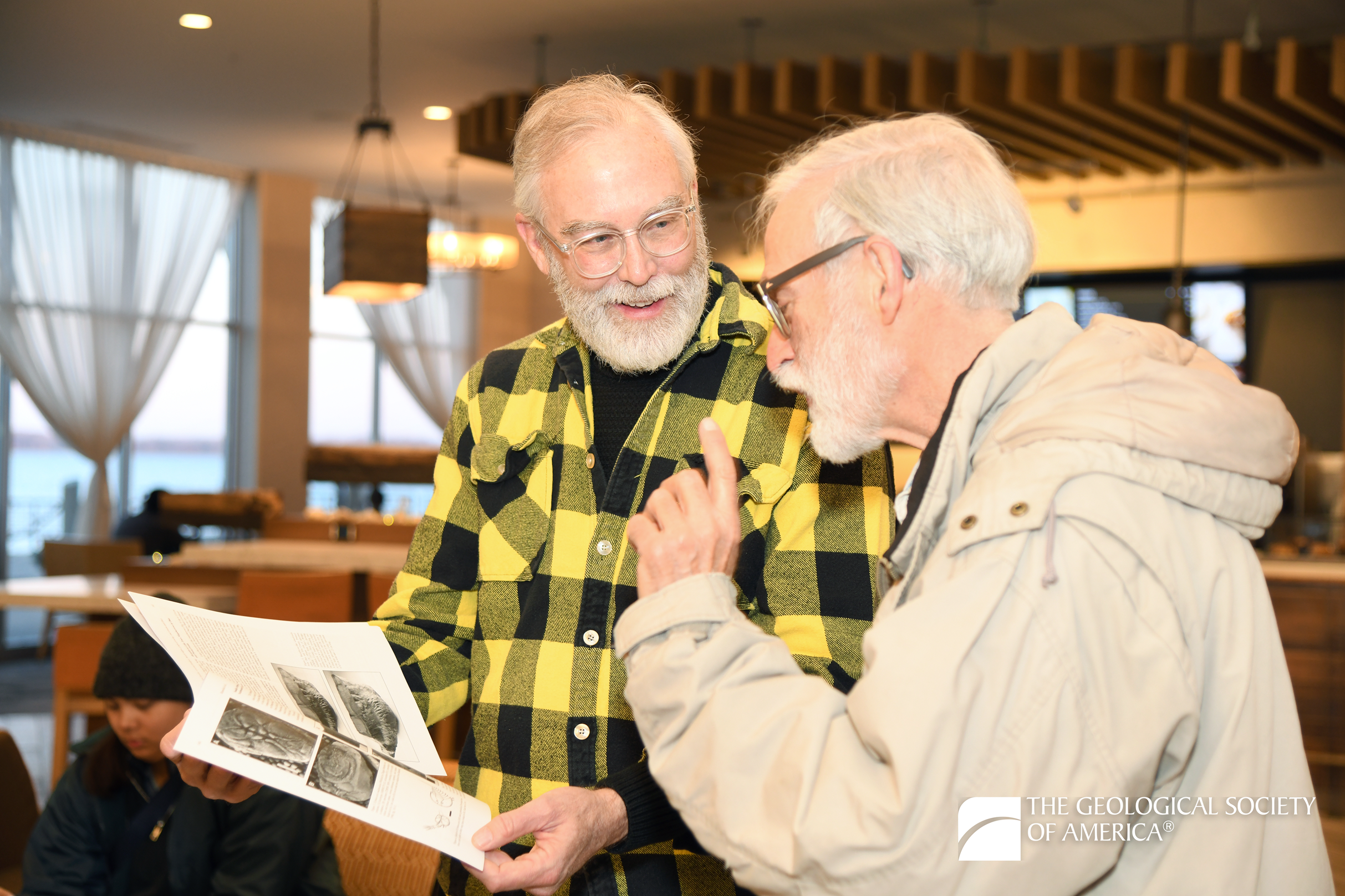 Two elderly men engage in discussion, examining geological documents. One wears a yellow plaid shirt, the other a beige jacket. The setting is a well-lit indoor space.