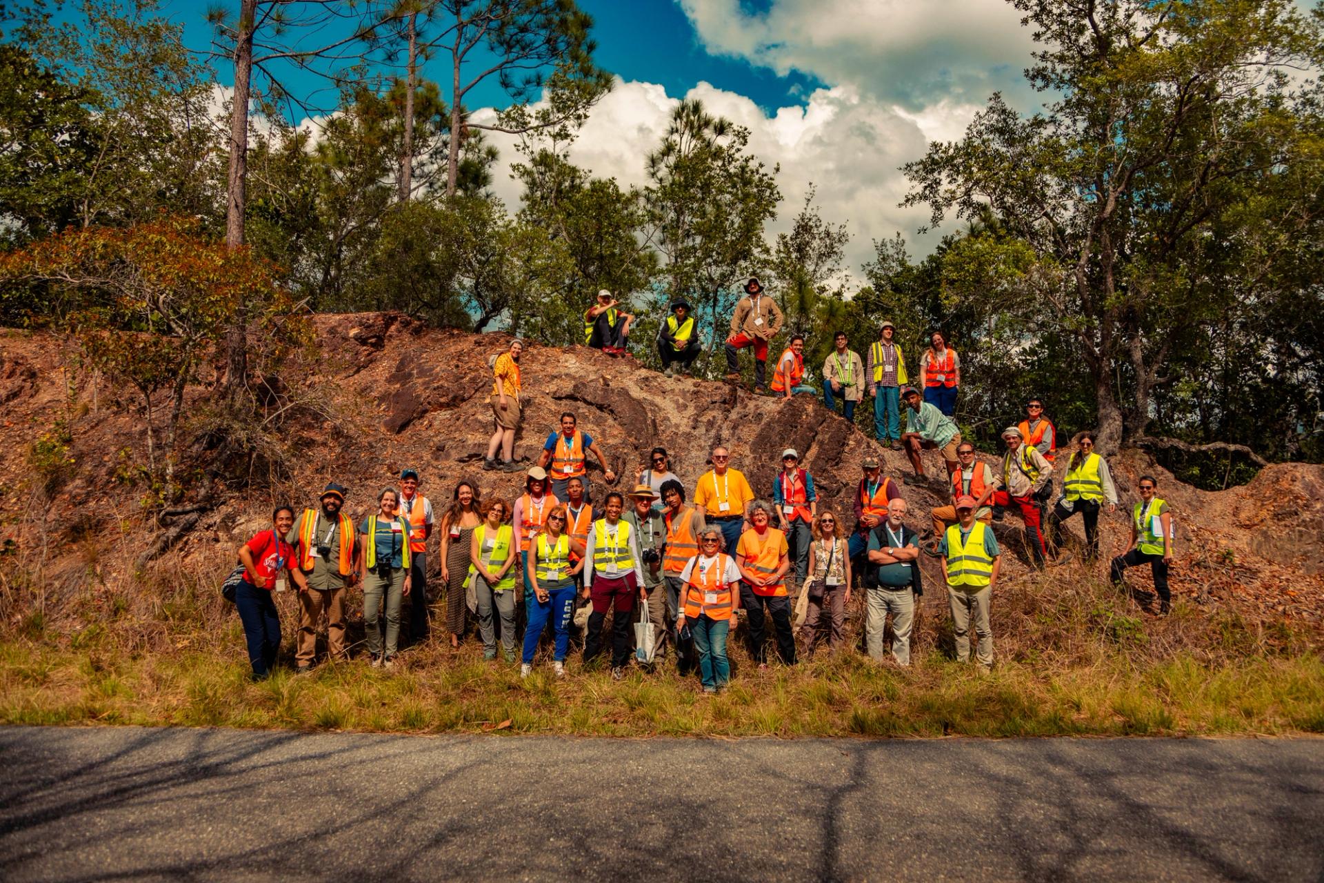 A large group of geologists and GSA Thompson Field Forum 2025 participants wearing safety vests and conference lanyards pose on and around a rocky outcrop surrounded by trees and vegetation during an outdoor field excursion in Cuba.