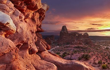 Sunset over Arches National Park, highlighting rugged red rock formations and a dramatic sky. The scene conveys a serene, majestic natural beauty.