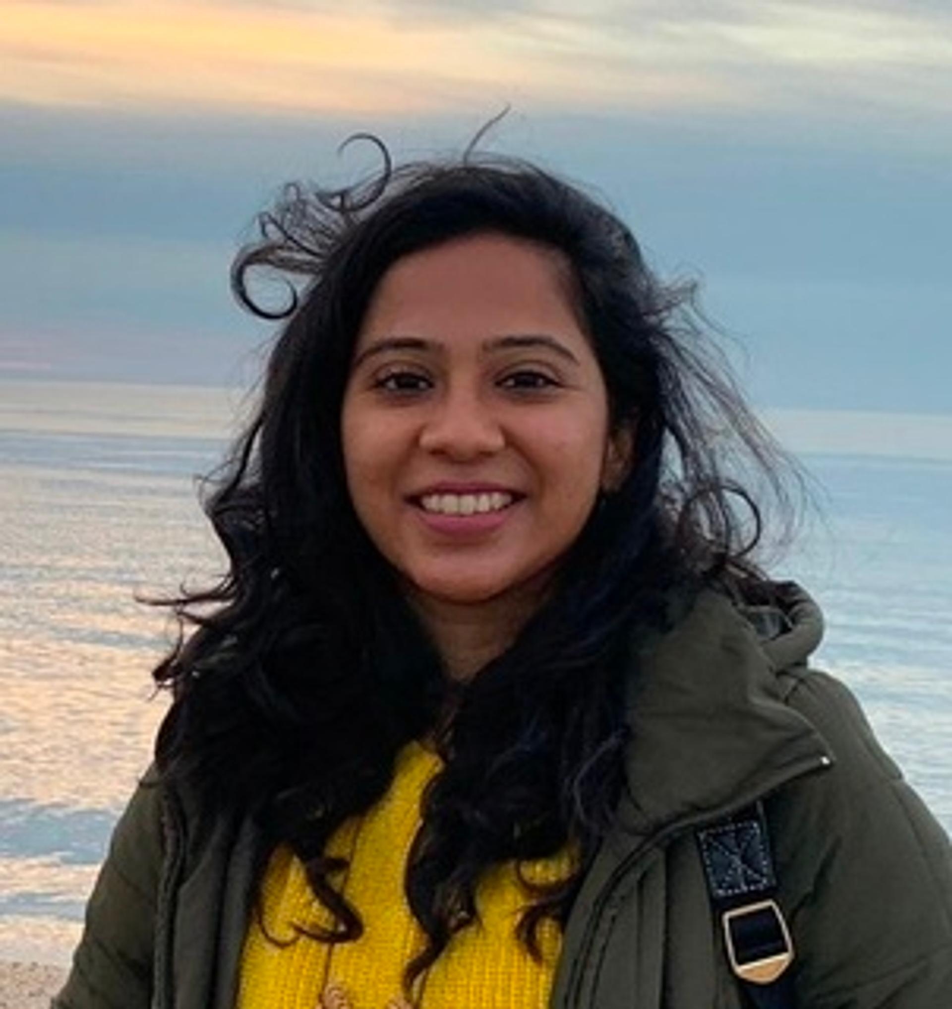 Urbi Basu in a green jacket, golden yellow sweater with blue water and a blue sky in the background.
