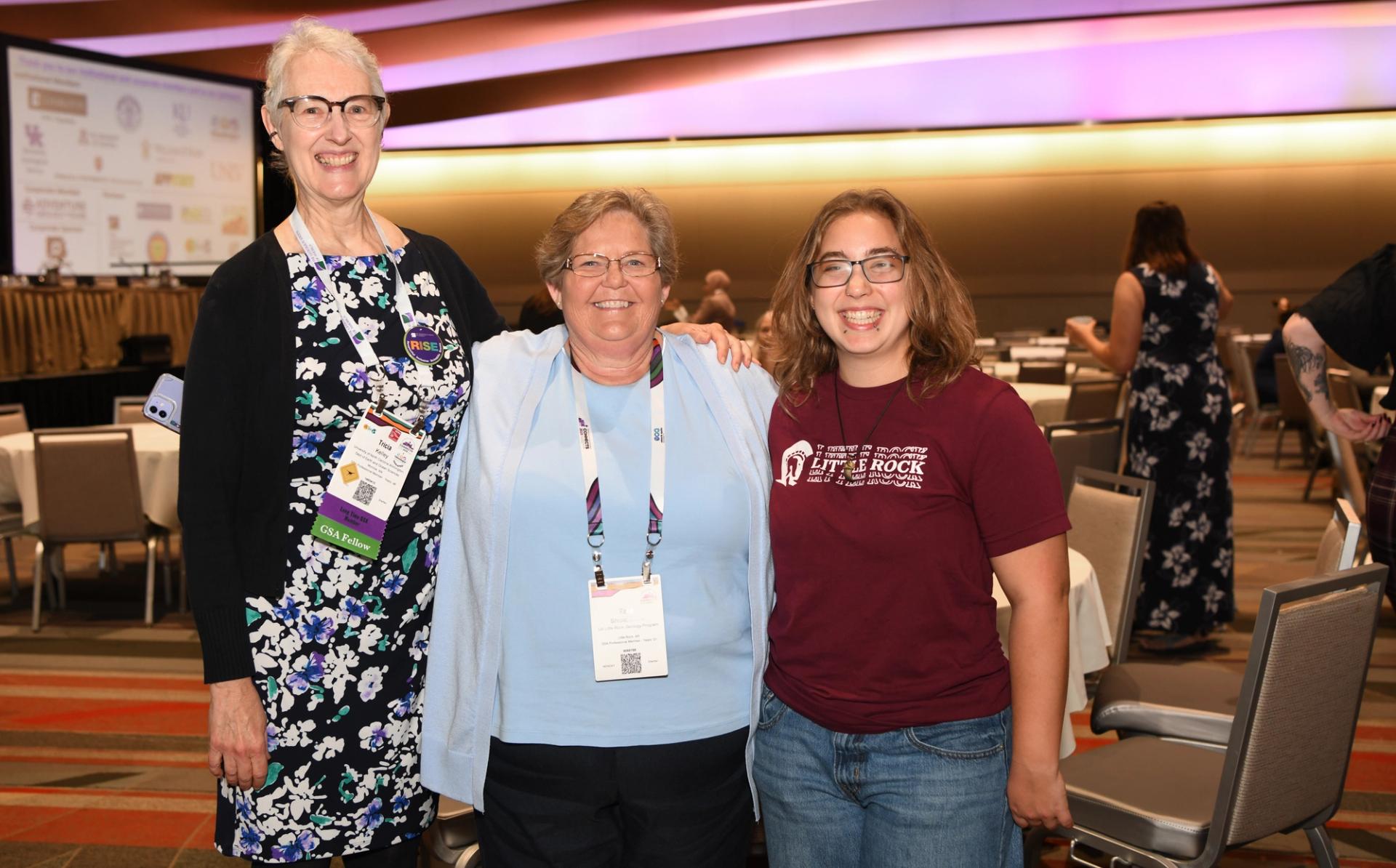 Three smiling female GSA Connects 2025 attendees of different generations pose together in a large conference ballroom, with one wearing a GSA Fellow badge ribbon and a RISE button, and a projected presentation screen visible in the background.
