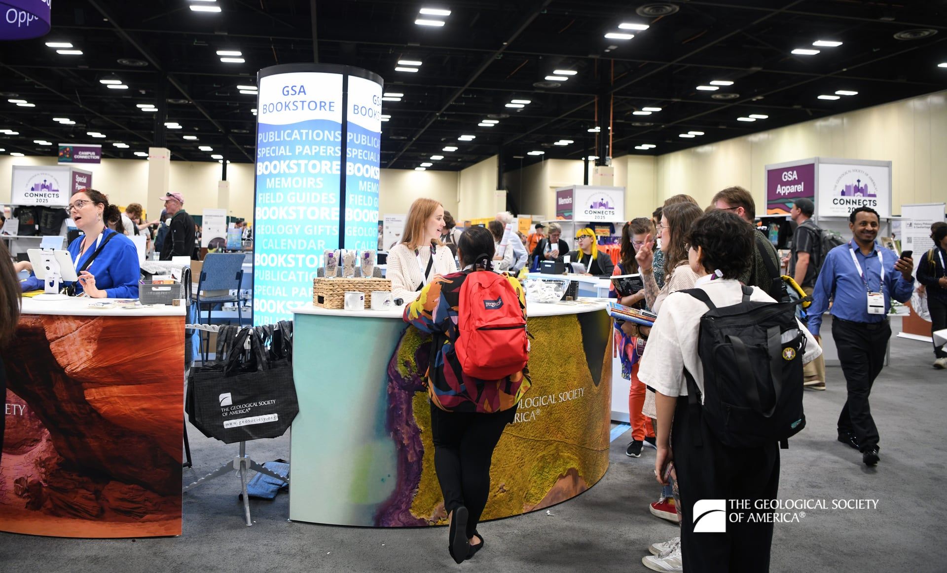 GSA Connects 2025 attendees gather around GSA Bookstore sales desks as GSA staff members ring up attendee purchases. The sales desks are covered in large, colorful photos of geologic features. 
