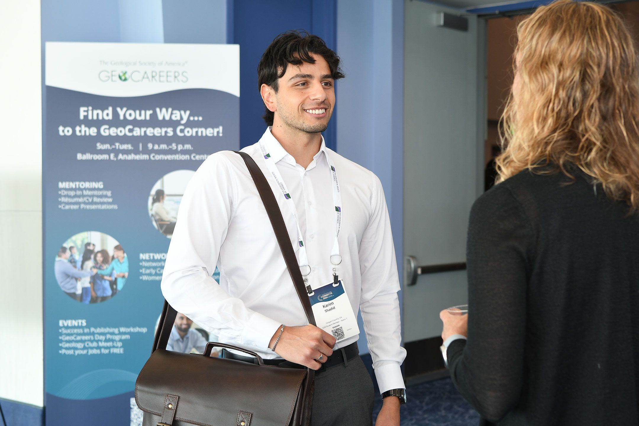 A conference attendee in business attire smiles at a person with their back to the camera.