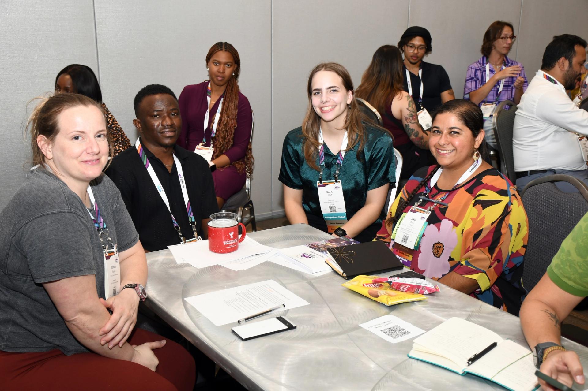 Four diverse GSA conference attendees wearing lanyards and badges smile together at a table during a professional development or networking session, with papers, notebooks, and conference materials spread across the table and other attendees visible in the background.