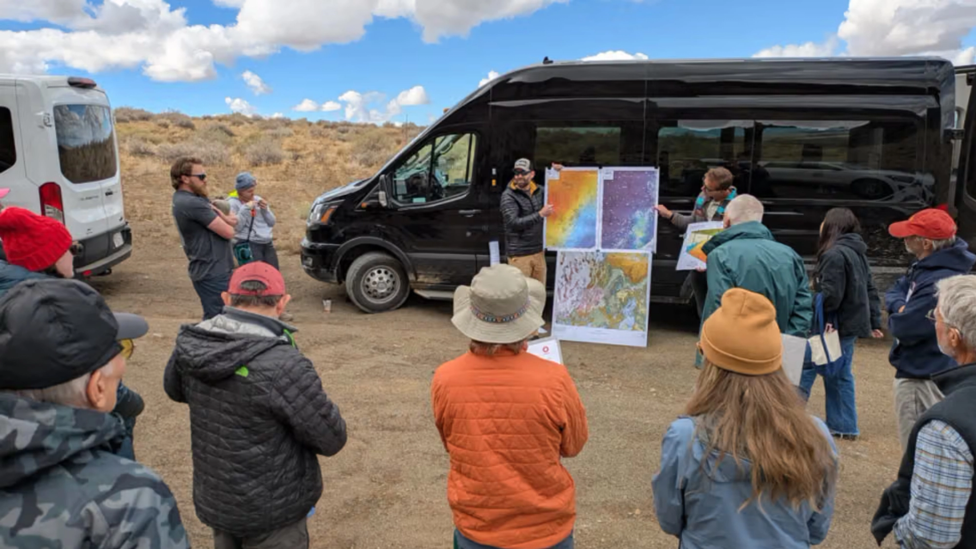 Field trip participants surrounding a speaker with a poster of three geologic maps in front of a black sprinter van.