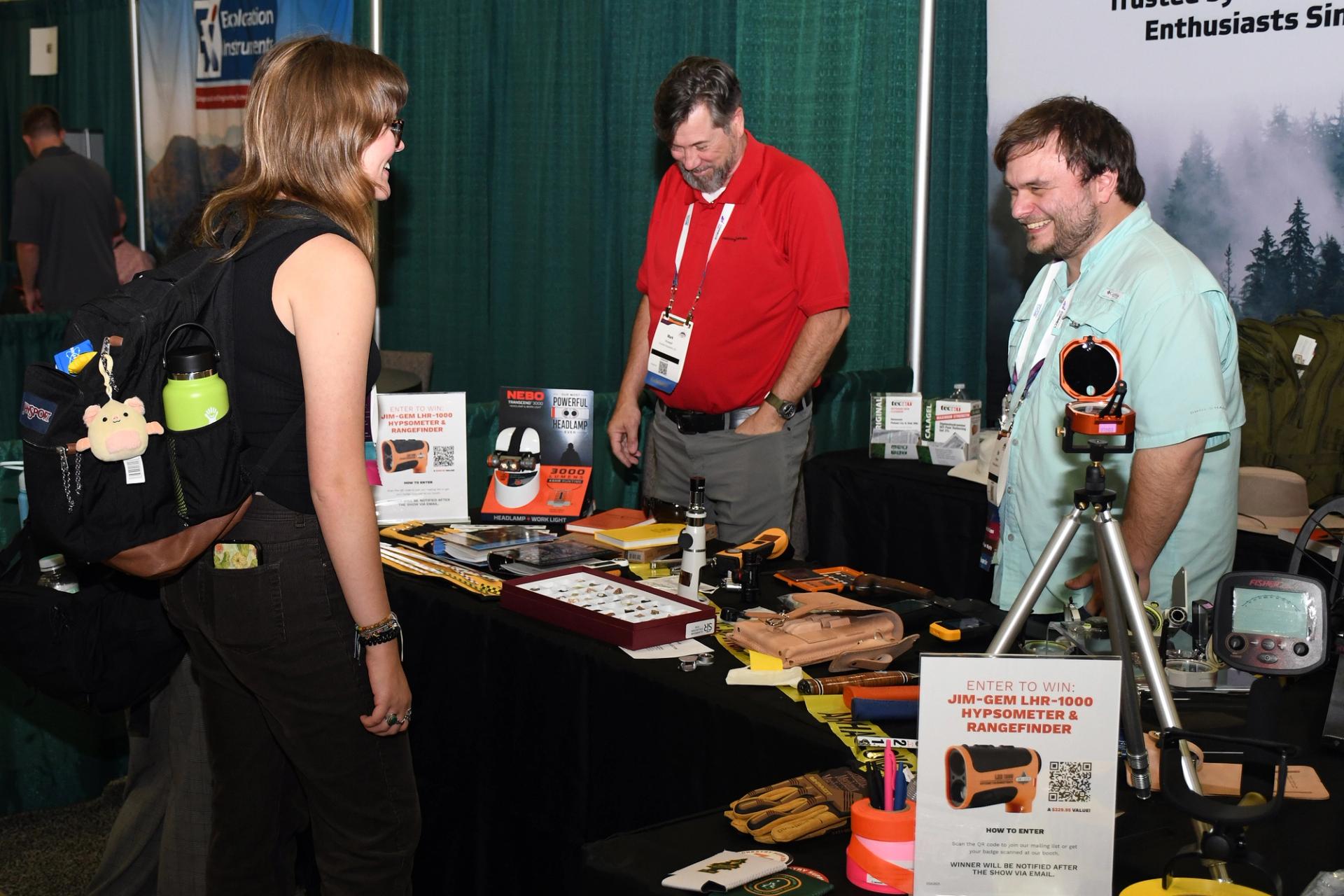 A GSA Connects attendee smiles while speaking with two exhibitors at a geoscience trade show booth displaying field equipment, tools, and instruments, with a giveaway sign visible in the foreground.