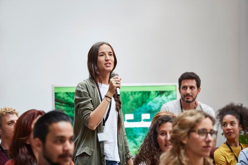 A young woman with long, dark hair in a braid, wearing a green jacket, holds a microphone and stands amid a group of seated audience members.