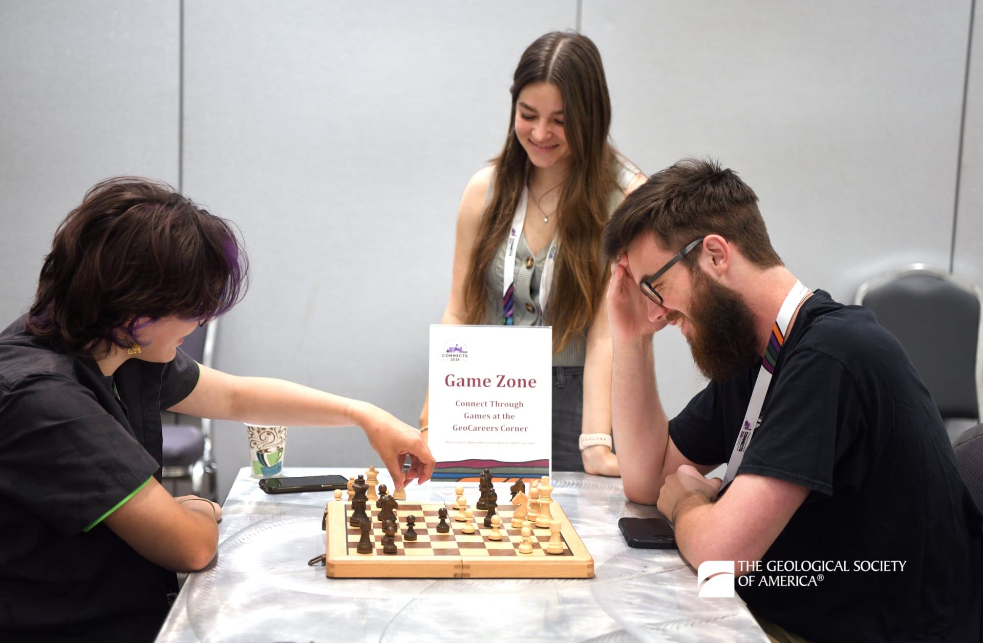 Two GSA Connects 2025 attendees are seated around a chess board and pieces in a Game Zone area as another attendee smiles, stands near the table, and watches the game.