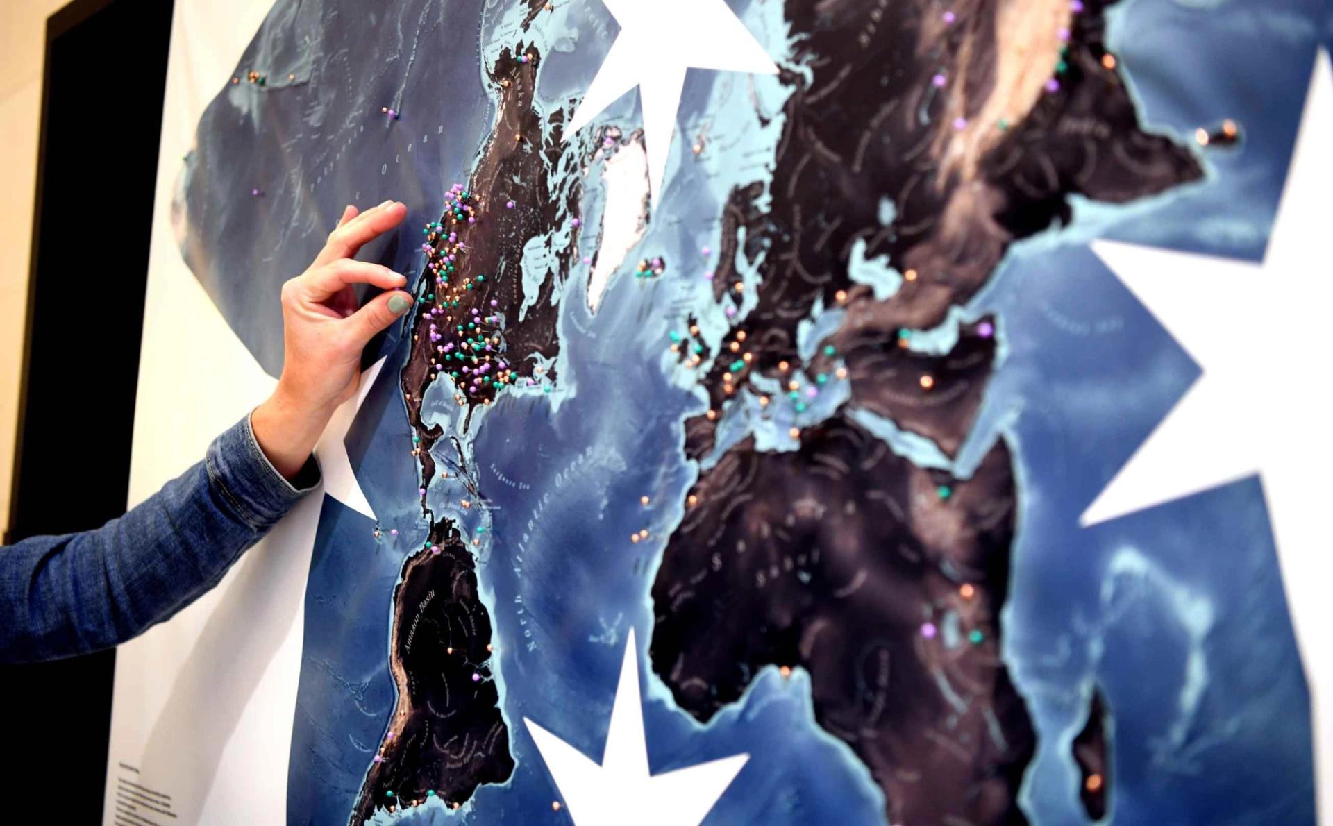 A close-up of a hand placing a colorful pin on a large stylized world map display covered in multicolored pins marking locations across the globe at GSA Connects 2025.