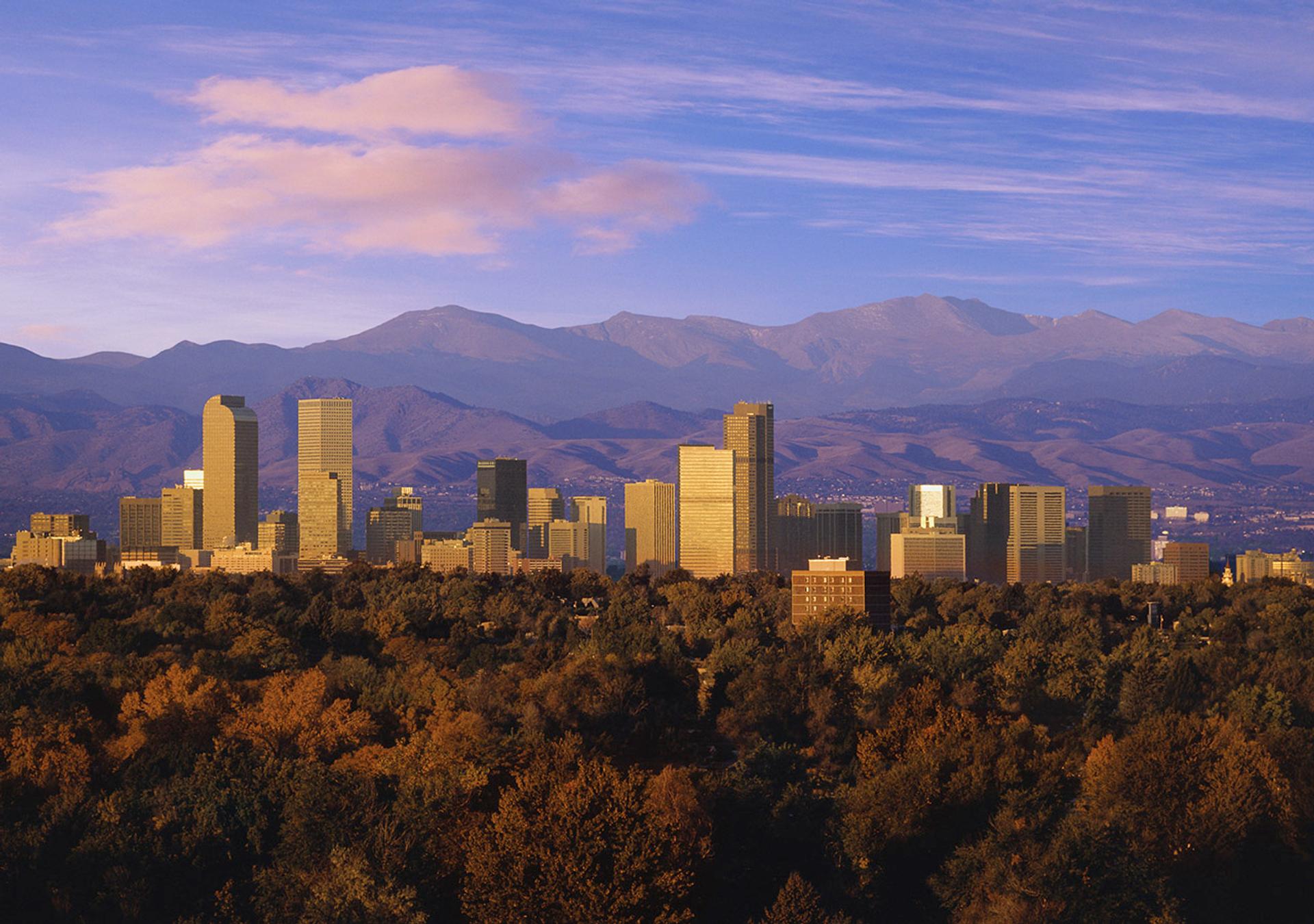Panoramic view of the Denver, Colorado skyline at dusk with Rocky Mountains in the background under a deep blue sky.