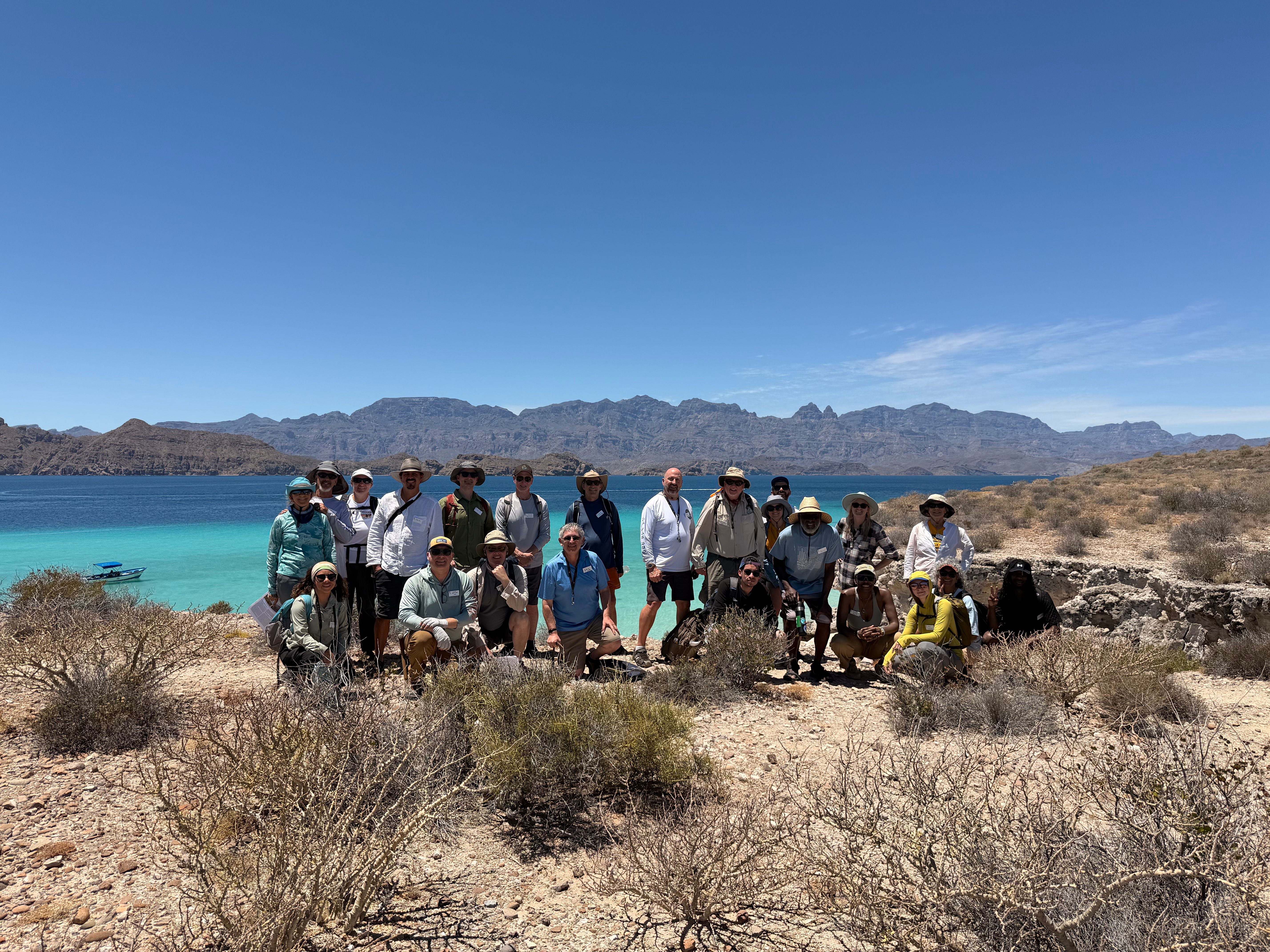 A large group of GSA Cordilleran Section Meeting 2026 field trip participants pose together on a rocky, arid coastline under a bright blue sky, with vivid turquoise water, a small boat, and dramatic mountain ridgelines visible in the background.