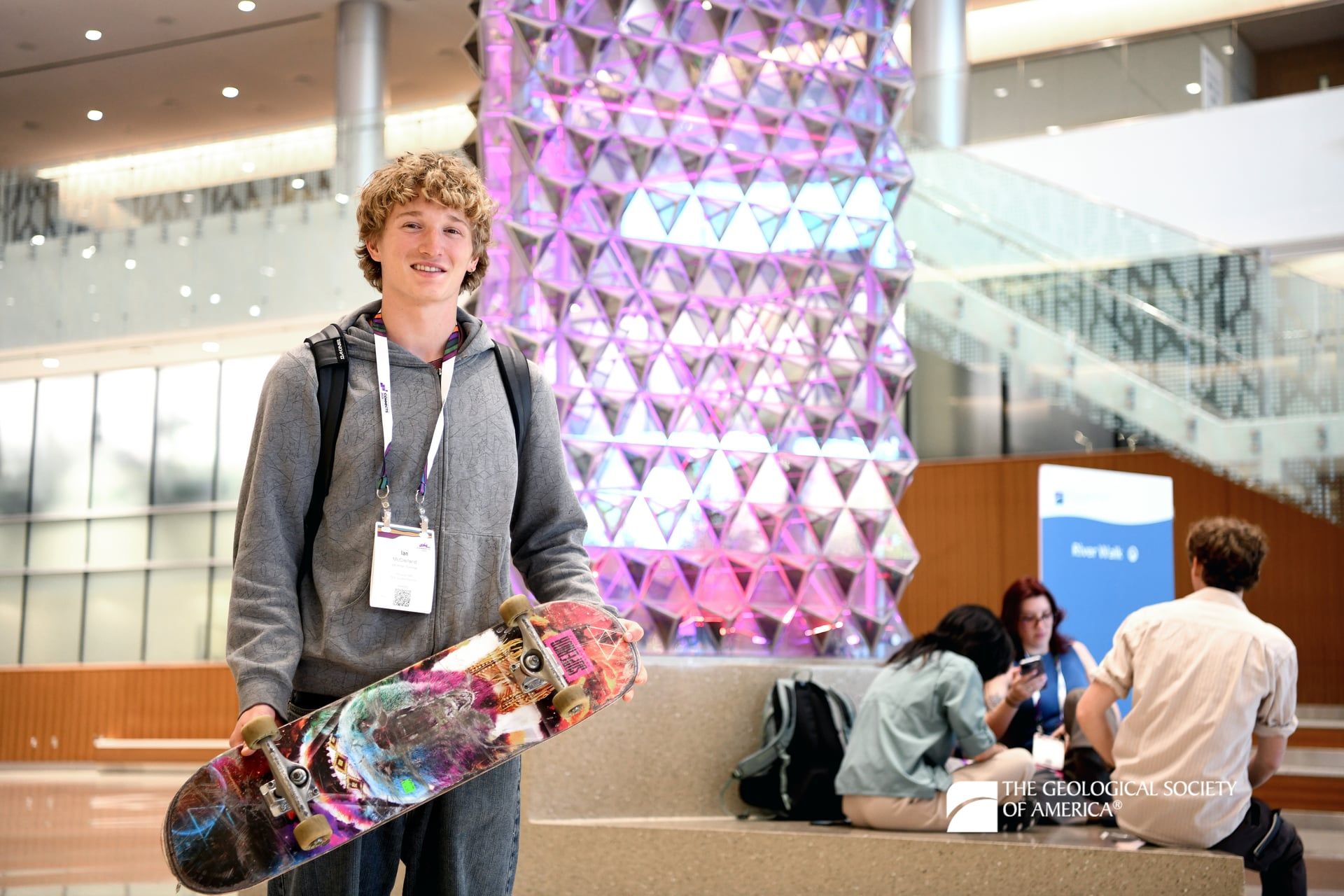 A GSA Connects 2025 attendee holds a brightly colored skateboard in front of a tall San Antonio Convention Center art statue. Meeting attendees in the background sit at the base of the sculpture.
