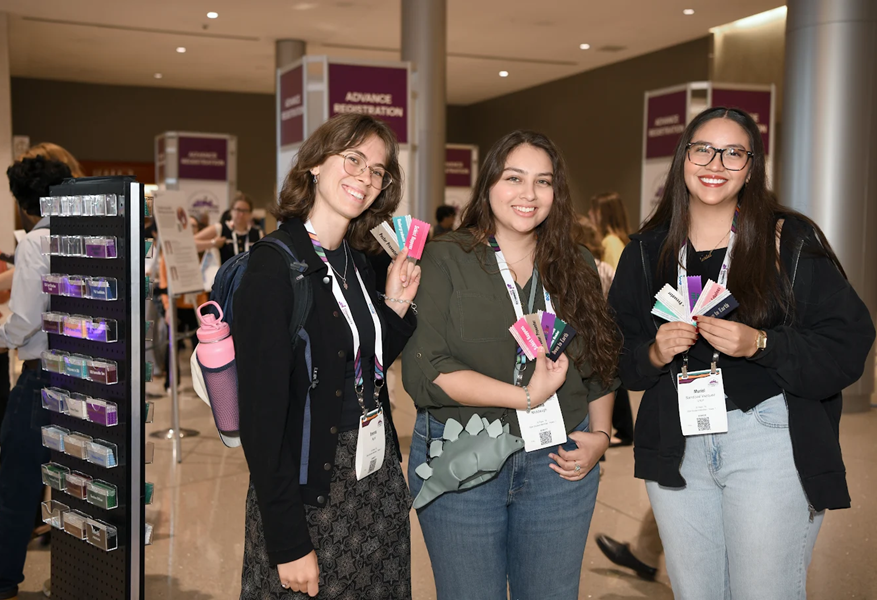 Three conference attendees pose with badge ribbons and smile near the registration area at GSA Connects 2025.