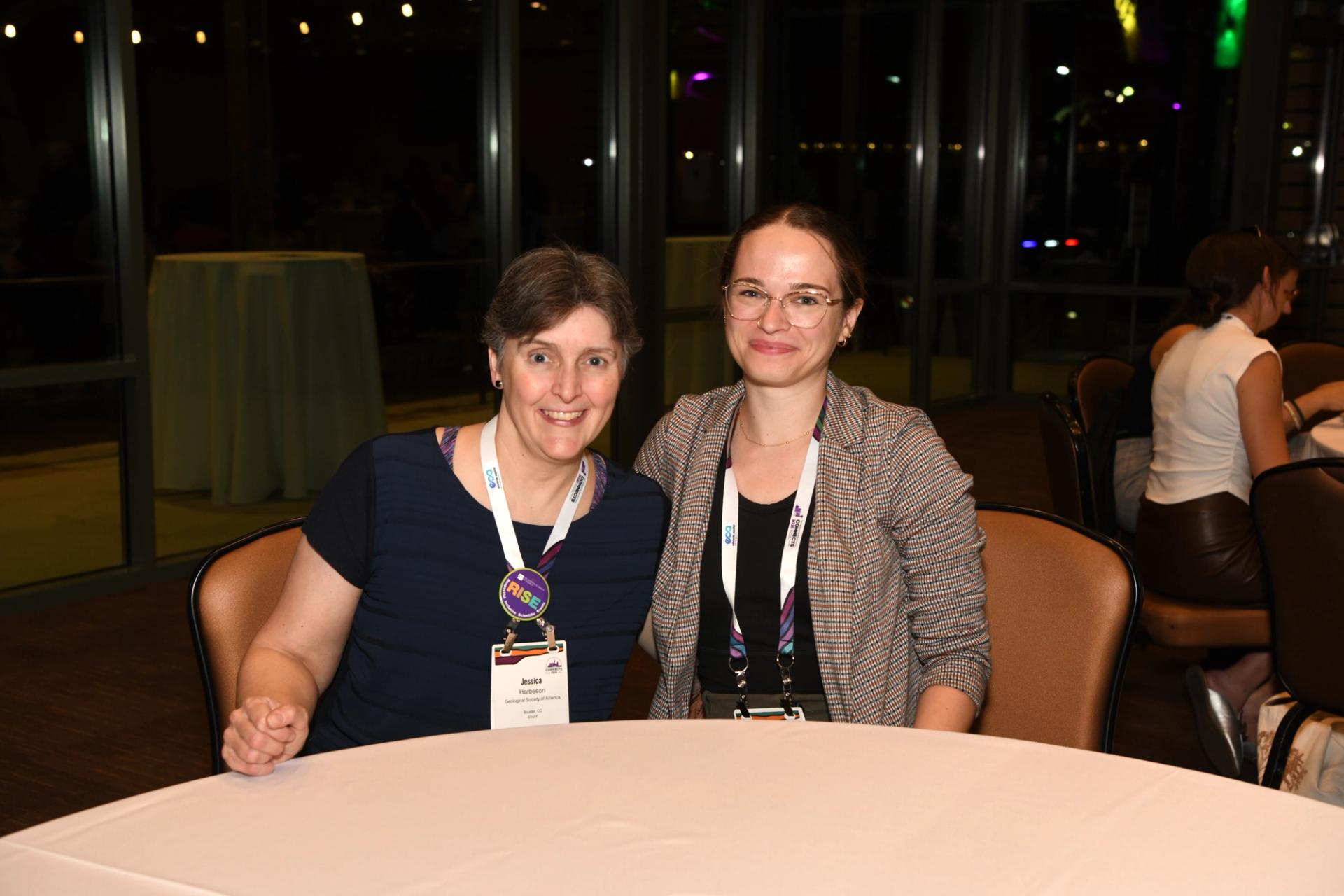 Two female GSA Staff wearing GSA Connects 2025 name badges and lanyards smile together at a round table during an evening networking event, Night at the Museum, with city lights and large windows visible in the background.