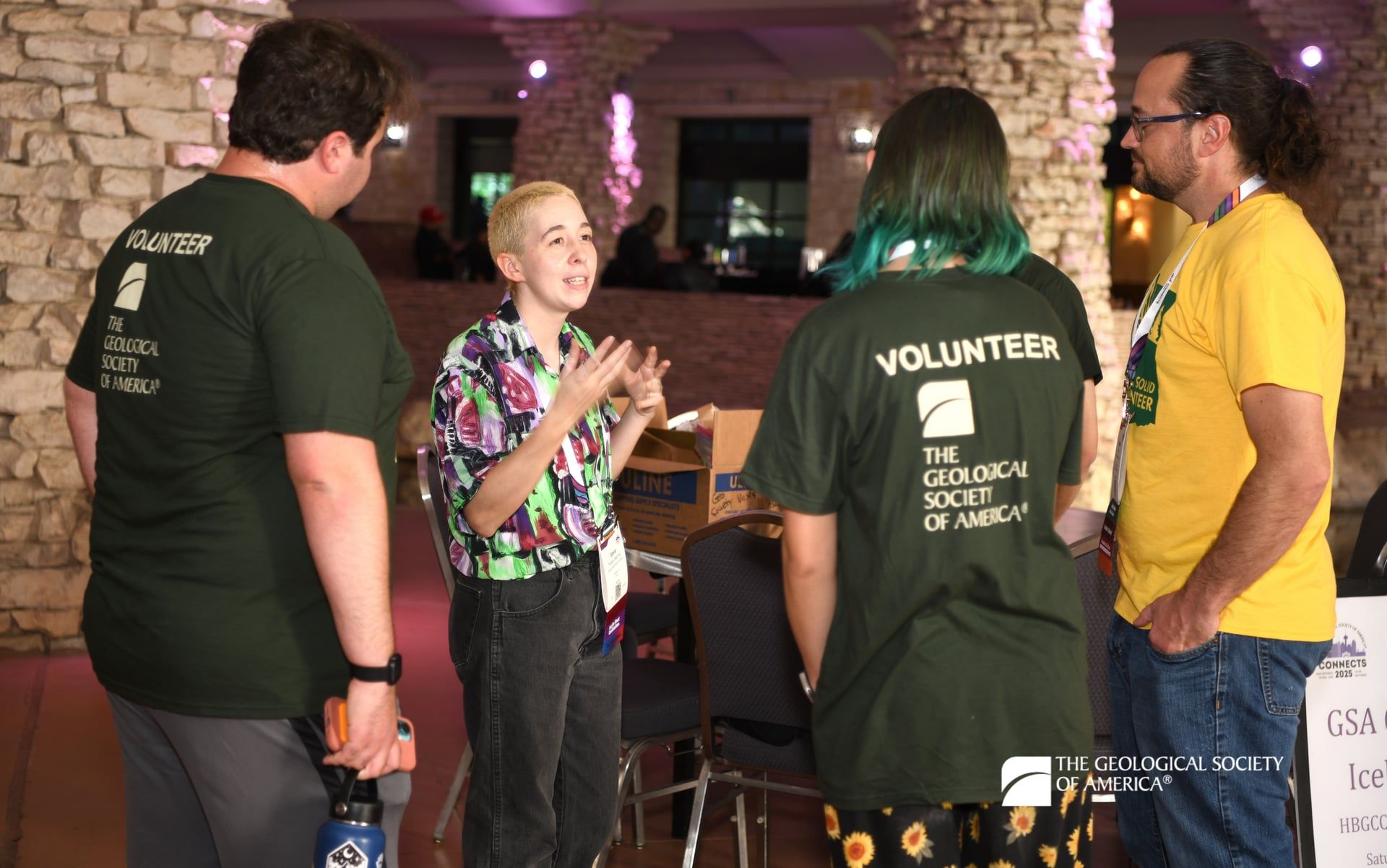 A GSA Connects staff member chats with three student volunteers in dressed in green and yellow volunteer t-shirts at a nighttime outdoor event. Behind them are stone pillars illuminated with purple uplighting.