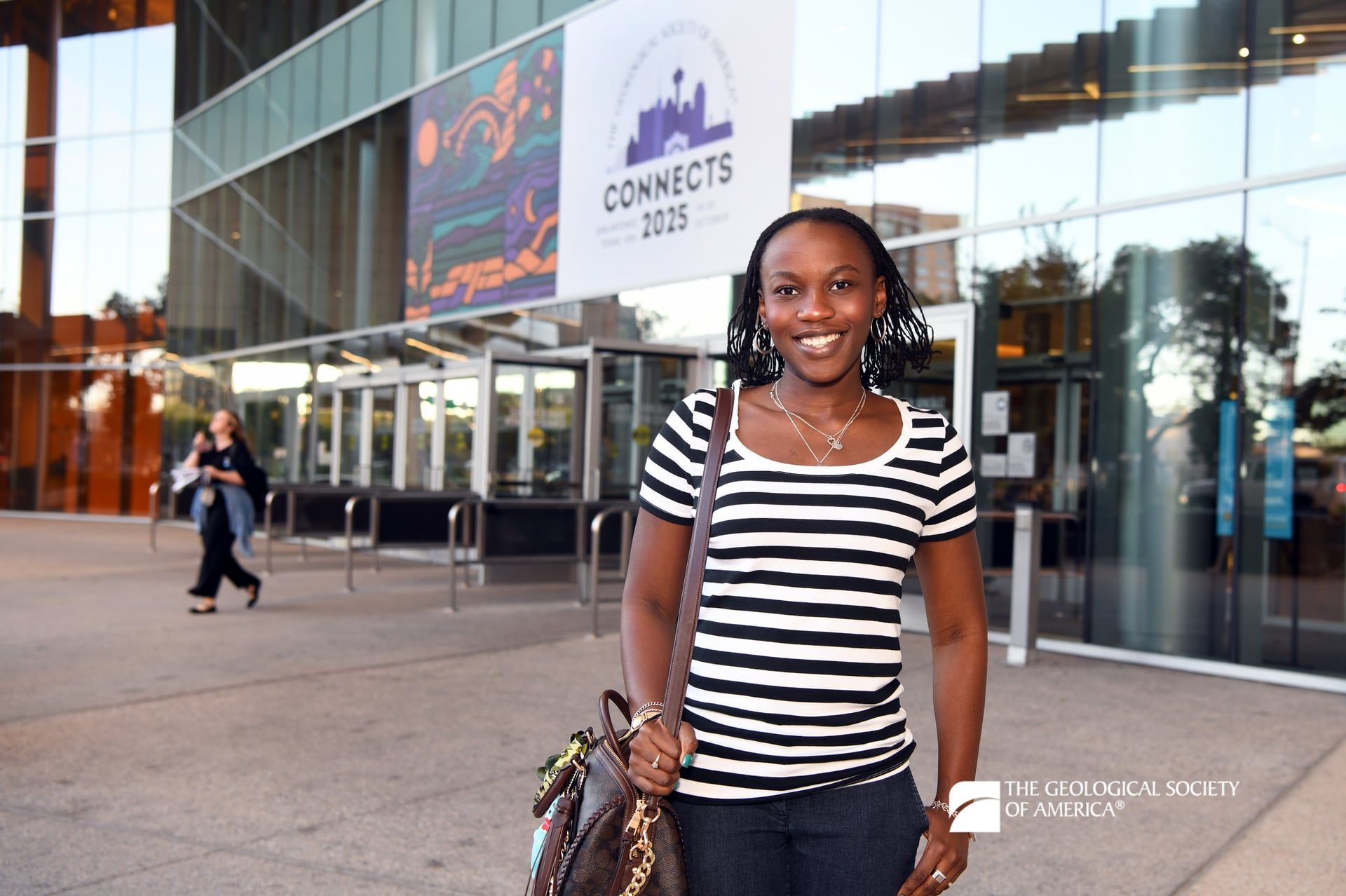 A GSA Connects 2025 attendee stands in front of a glass wall at the entrance of the San Antonio Convention Center. A large GSA Connects sign affixed to the building is in the background and displays above them.