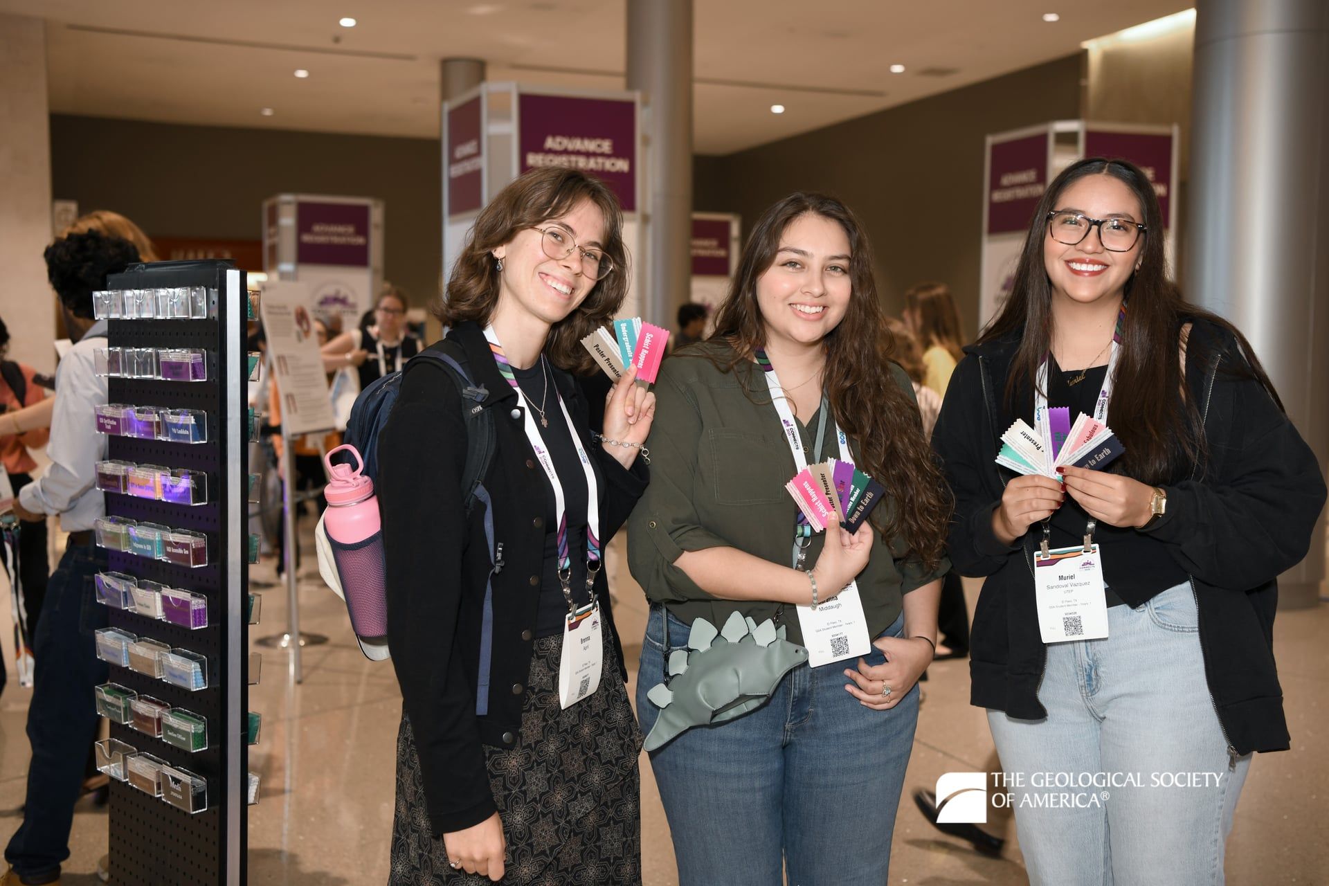 Three smiling GSA Connects 2025 attendees hold a variety of colorful badge ribbons as they stand in front of the Registration area of the meeting.