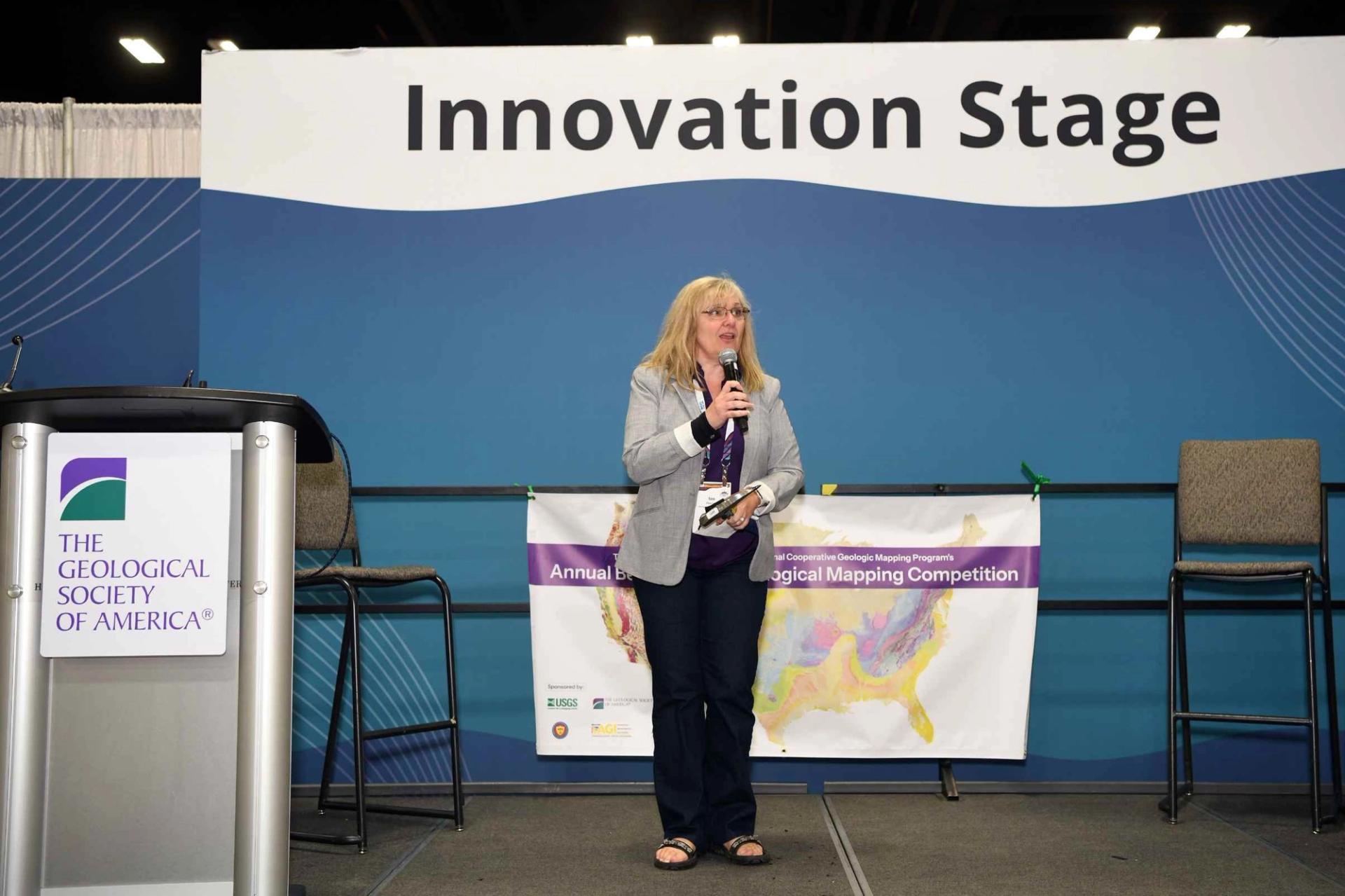 A female presenter holds a microphone and speaks on the GSA Innovation Stage in the Exhibit Hall, with a large Innovation Stage banner visible behind her, a Geological Society of America podium to her left, and a National Cooperative Geologic Mapping Program's Annual Best Geologic Mapping Competition banner in the background.