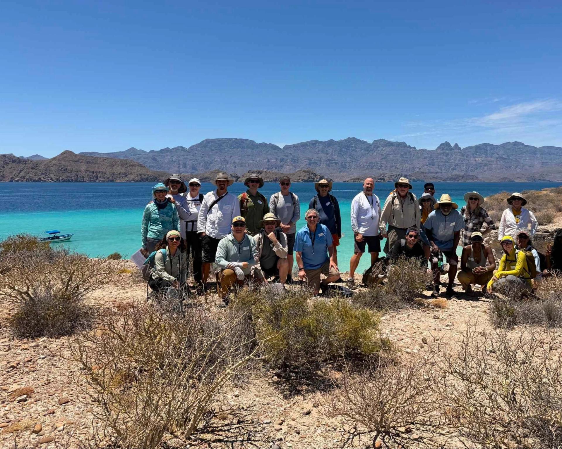 A large group of GSA Cordilleran Section Meeting 2026 field trip participants pose together on a rocky, arid coastline under a bright blue sky, with vivid turquoise water, a small boat, and dramatic mountain ridgelines visible in the background.