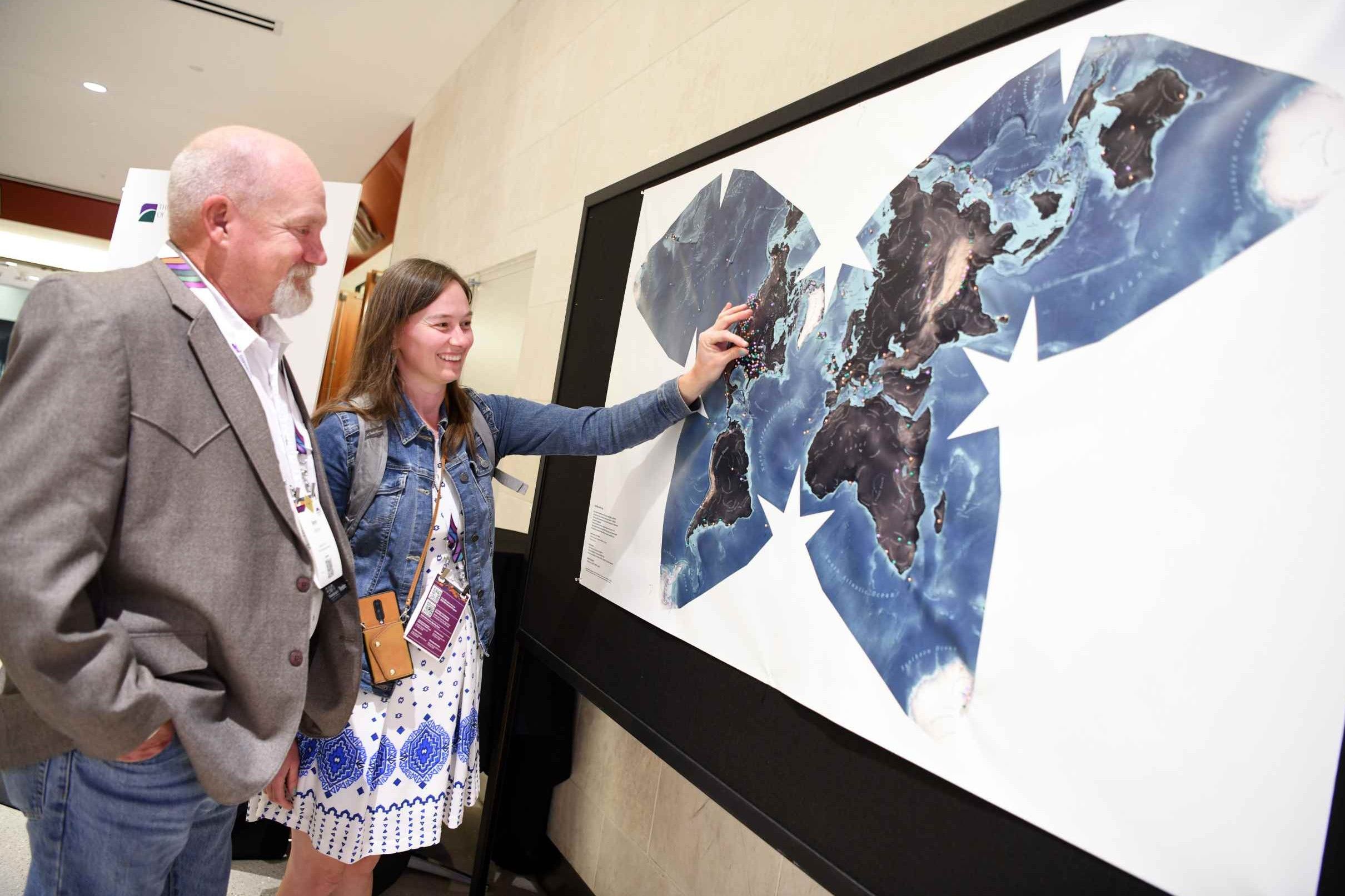 Two GSA Connects 2025 attendees wearing name badges examine a large world map display with pinned locations that attendees have pinned based on where they are from, with a young woman pointing to a location on the map while an older man looks on with interest.