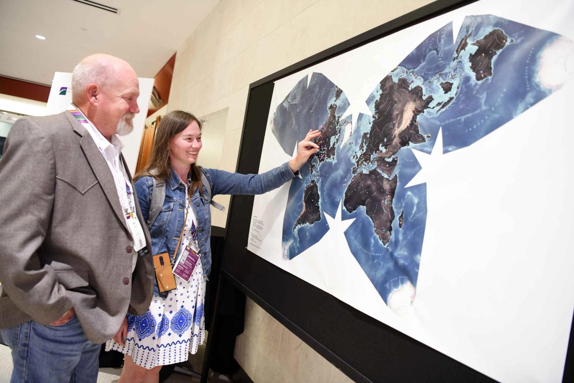 Two GSA Connects 2025 attendees wearing name badges examine a large world map display with pinned locations that attendees have pinned based on where they are from, with a young woman pointing to a location on the map while an older man looks on with interest.