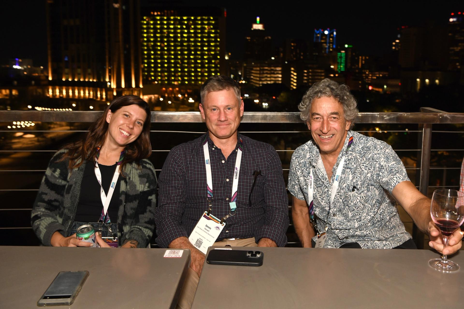 Three smiling GSA Connects 2025 attendees wearing lanyards and badges relax together at an outdoor rooftop bar during an evening networking event, with a glowing San Antonio city skyline visible behind them.