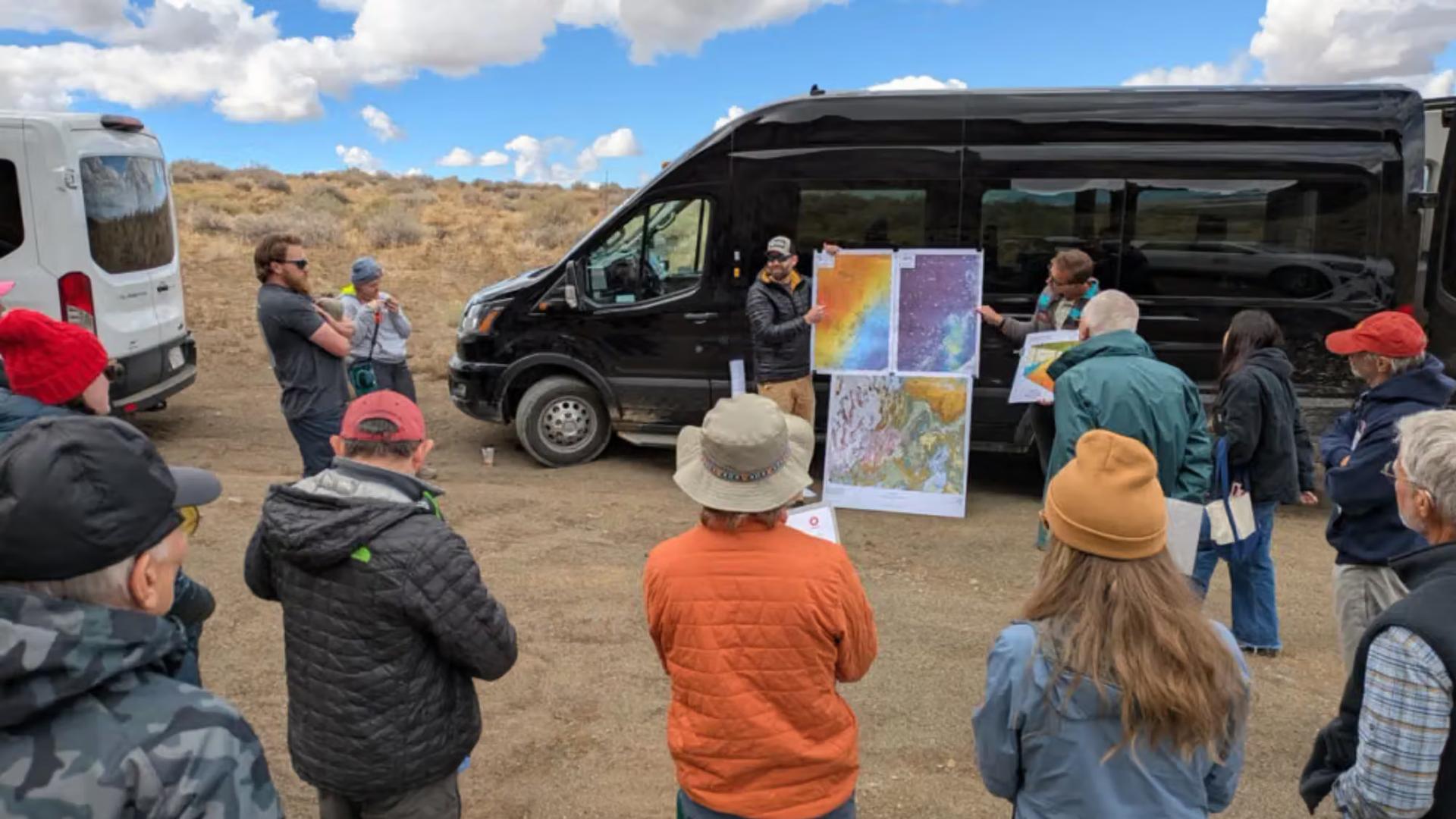 Field trip participants surrounding a speaker with a poster of three geologic maps in front of a black sprinter van.