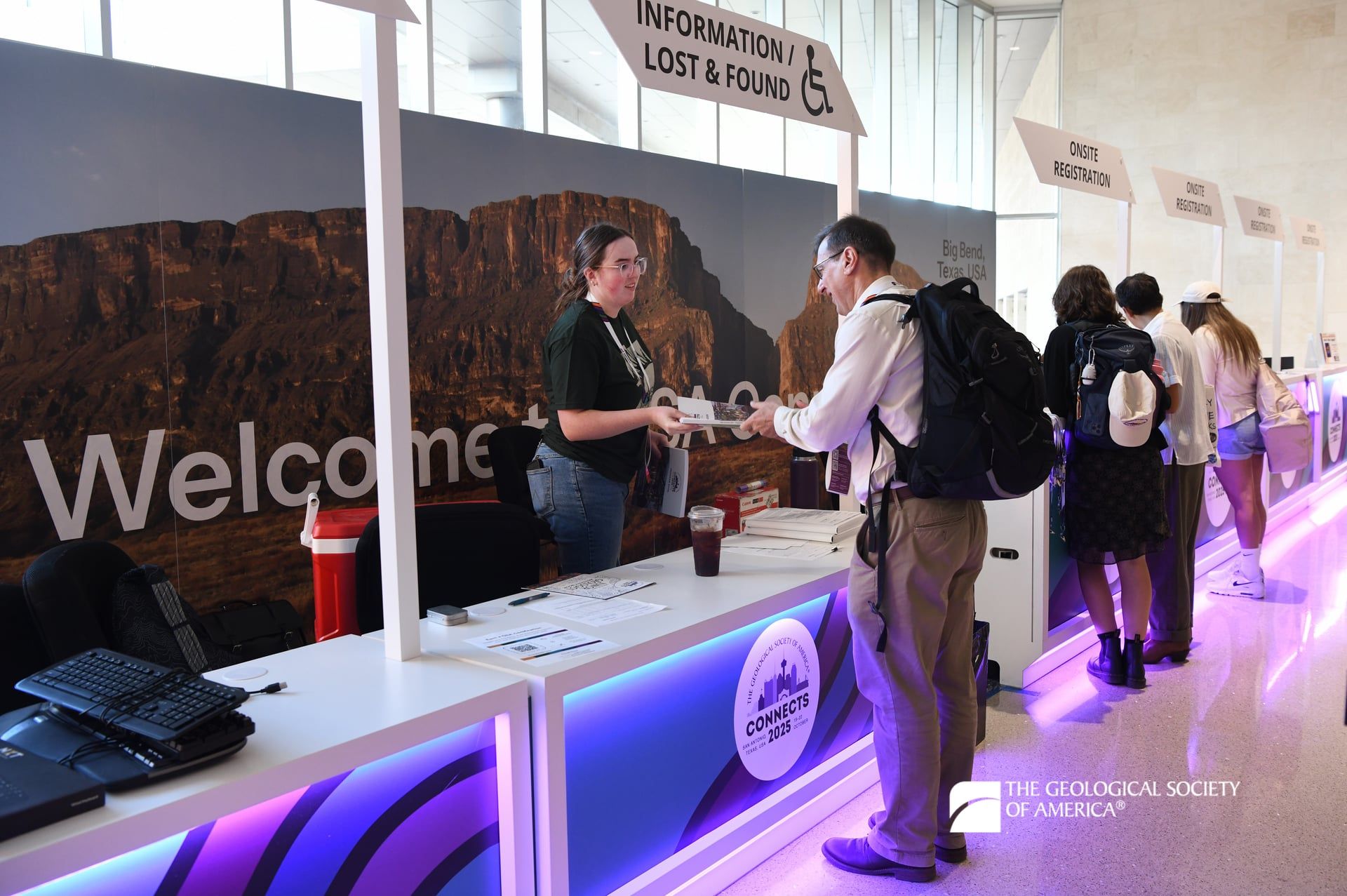 A GSA Connects 2025 student volunteer provides a program book to an attendee at the Registration desk. Behind them is a large backdrop featuring an image of geology in Texas. Meeting registration desks are illuminated with purple lights.