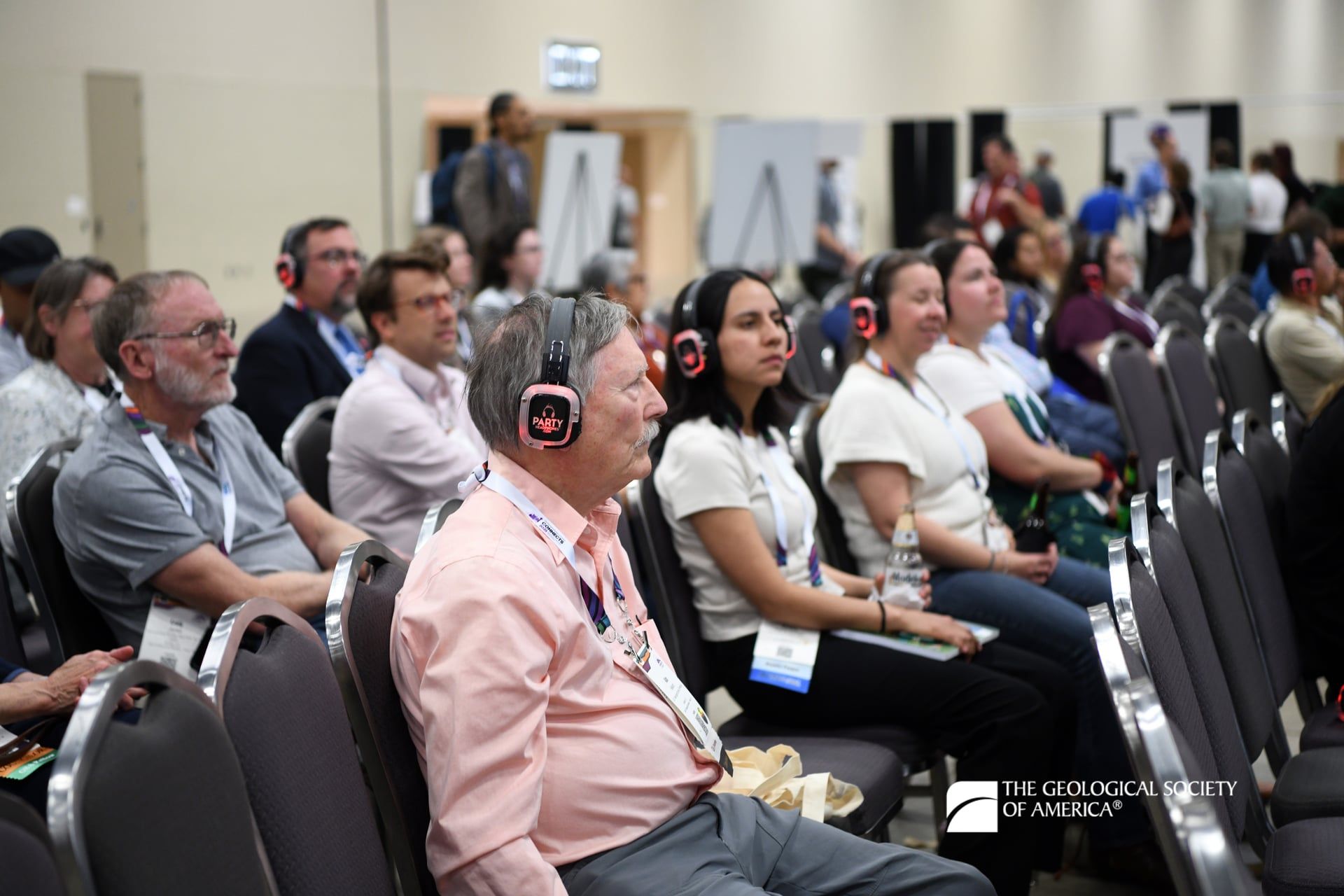 A group of GSA Connects 2025 attendees sit in the audience section of the Innovation Stage. Some wear headphones with glowing red earpieces.