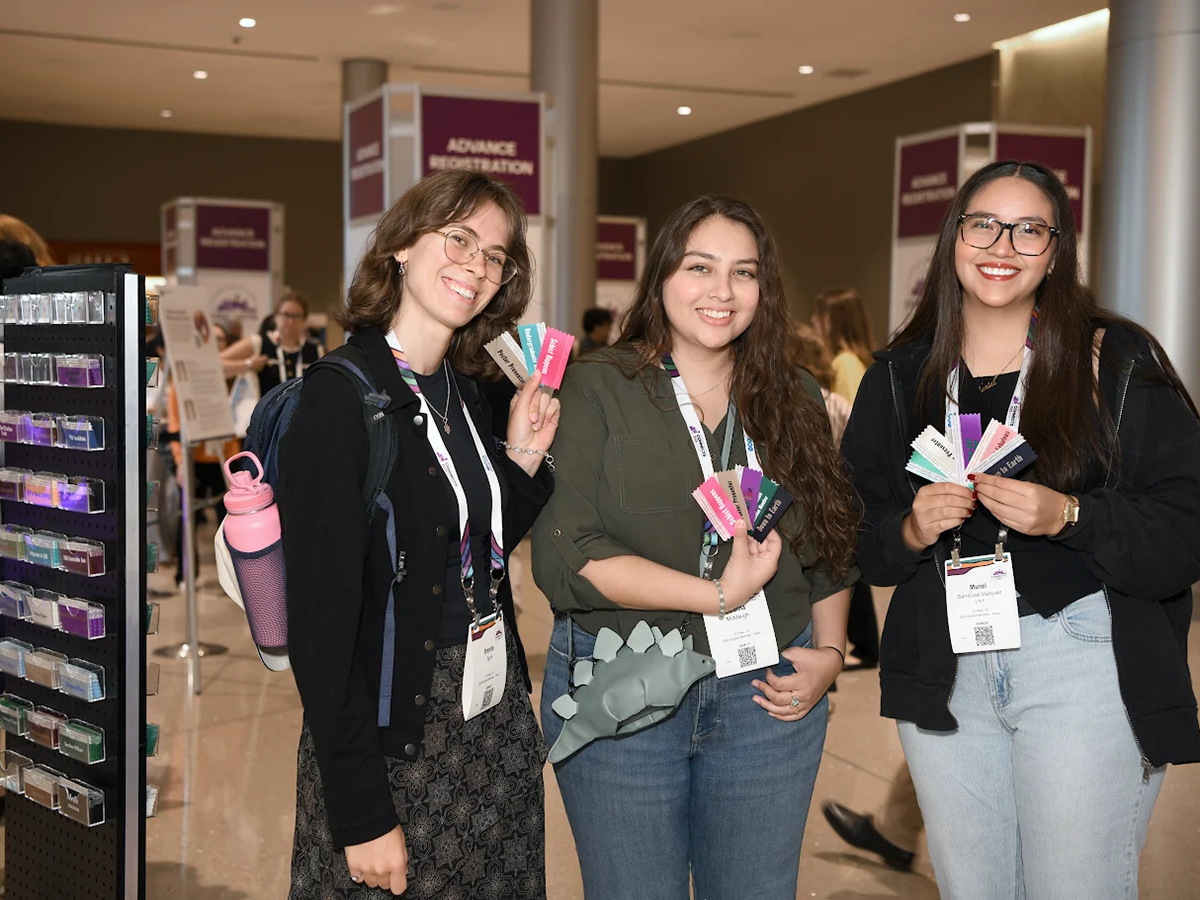 Three conference attendees pose with badge ribbons and smile near the registration area at GSA Connects 2025.