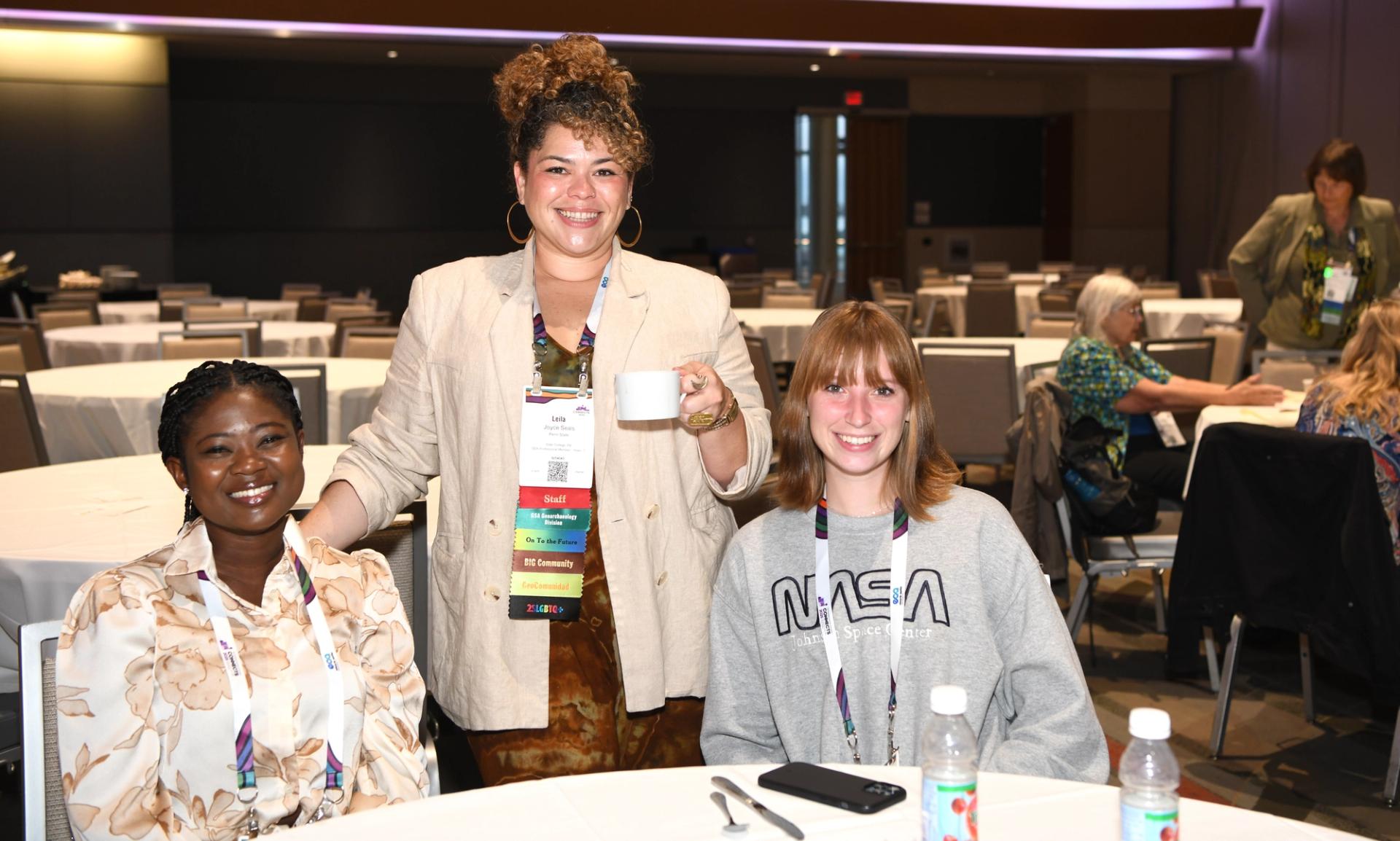 Three smiling GSA Connects 2025 attendees pose together at a round table during a networking event, one standing and holding a coffee cup while the other two are seated, with multiple colorful badge ribbons visible.