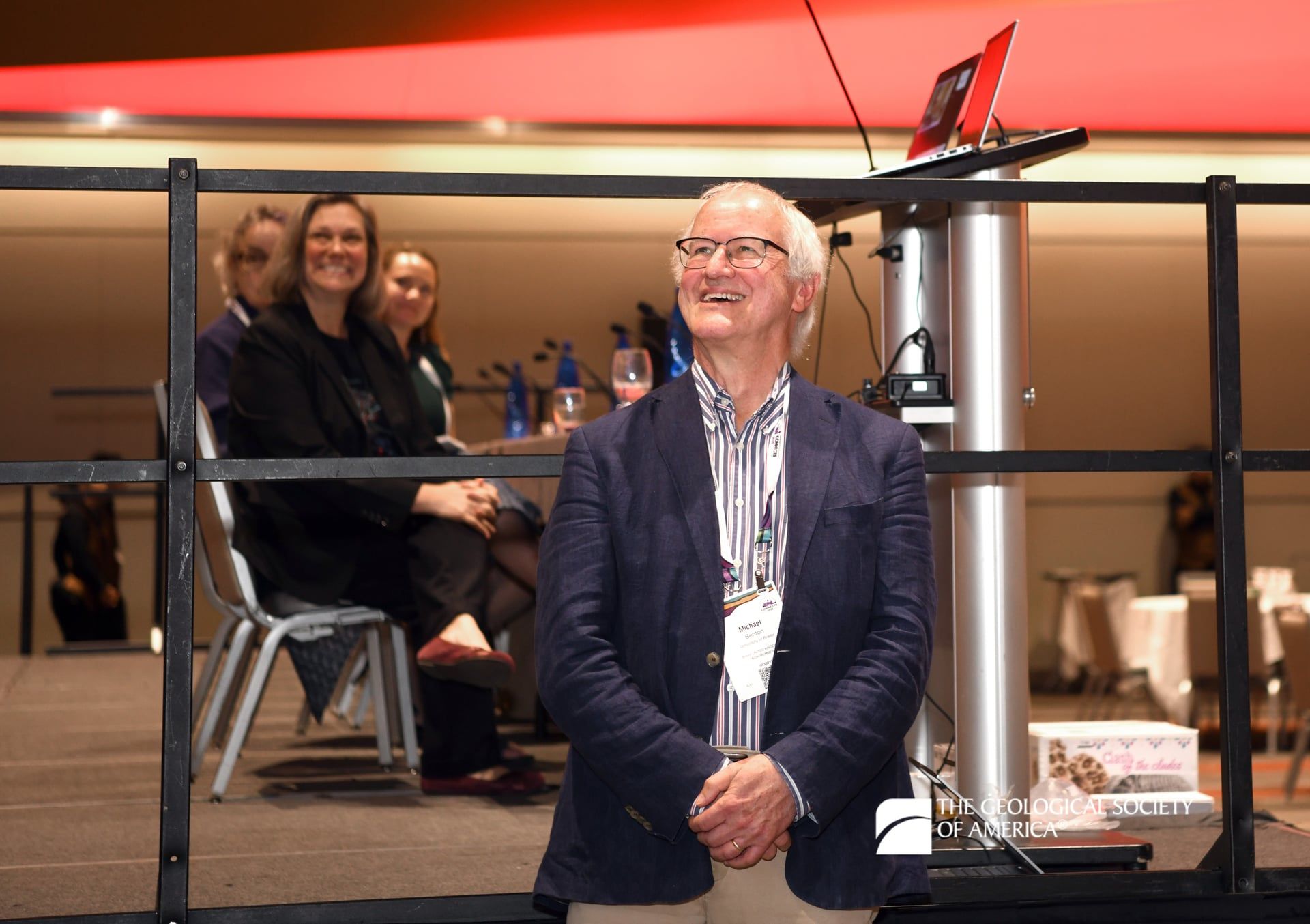 A GSA Connects 2025 attendee beams with a smile as they stand near a meeting stage. Three attendees in the background sit in chairs on a platform.