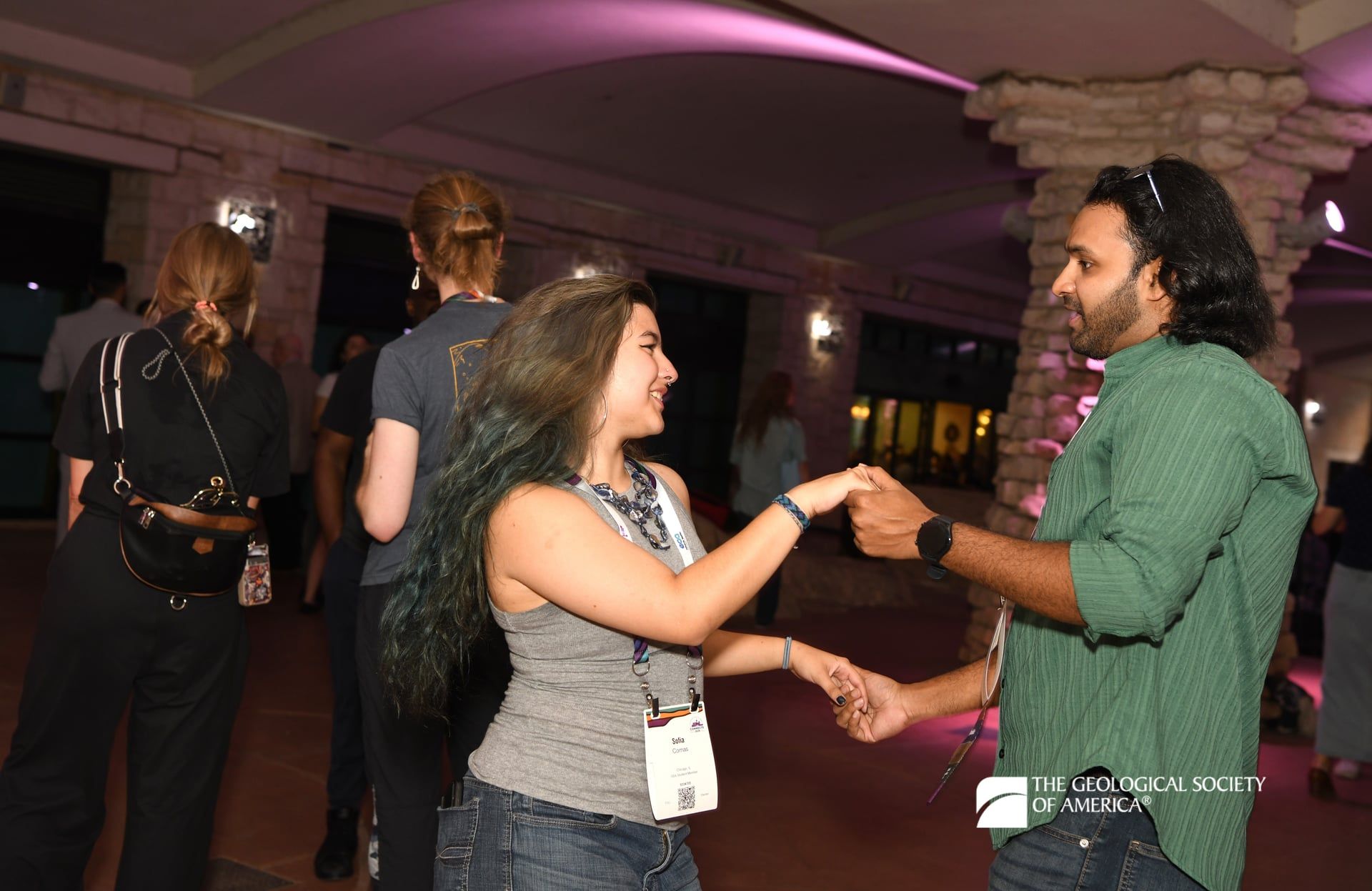 Two GSA Connects 2025 attendees dance at the outdoor icebreaker event. Additional attendees stand in the background amongst the patio's stone pillars that are lit with purple uplights.