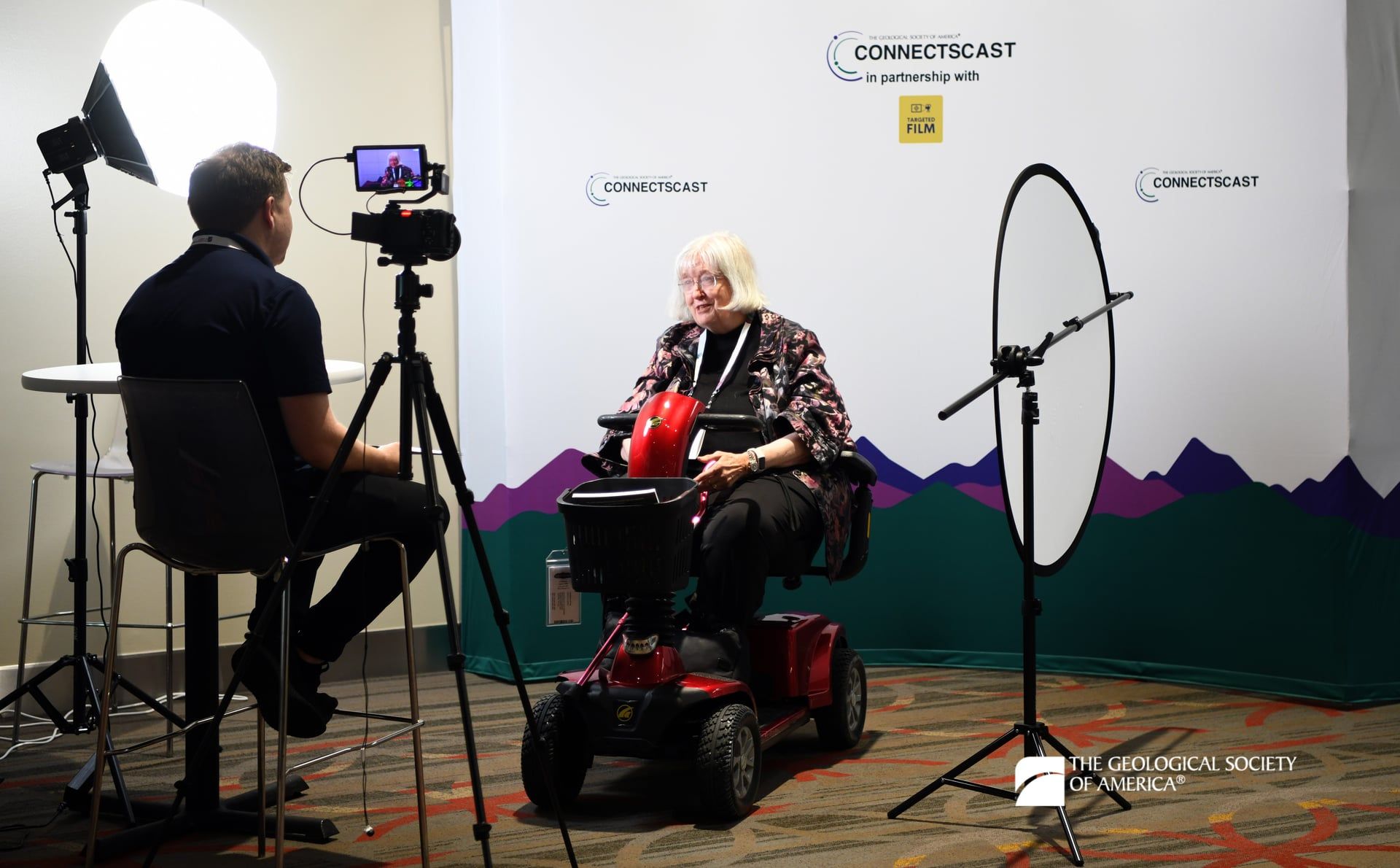 A GSA Connects 2025 attendee sits in a red motorized scooter as they are interviewed. Behind them is a white backdrop accented at the bottom by a green mountain range. Various lighting equipment and the interviewer is in the foreground.