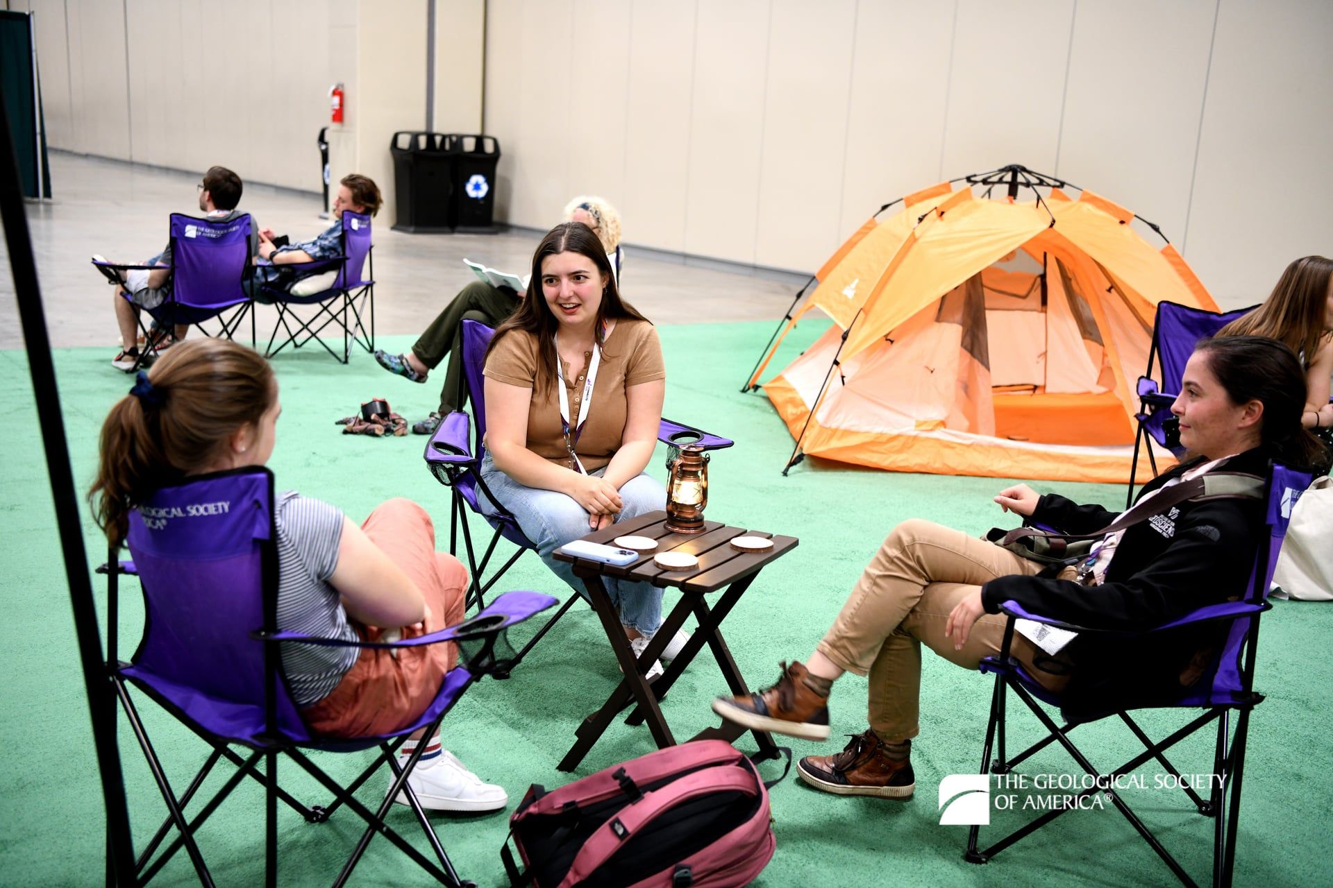 Seven GSA Connects 2025 attendees sit in purple camp chairs, some around tables, and relax while chatting with each other at the meeting. An orange tent has been placed atop green carpet in the background.
