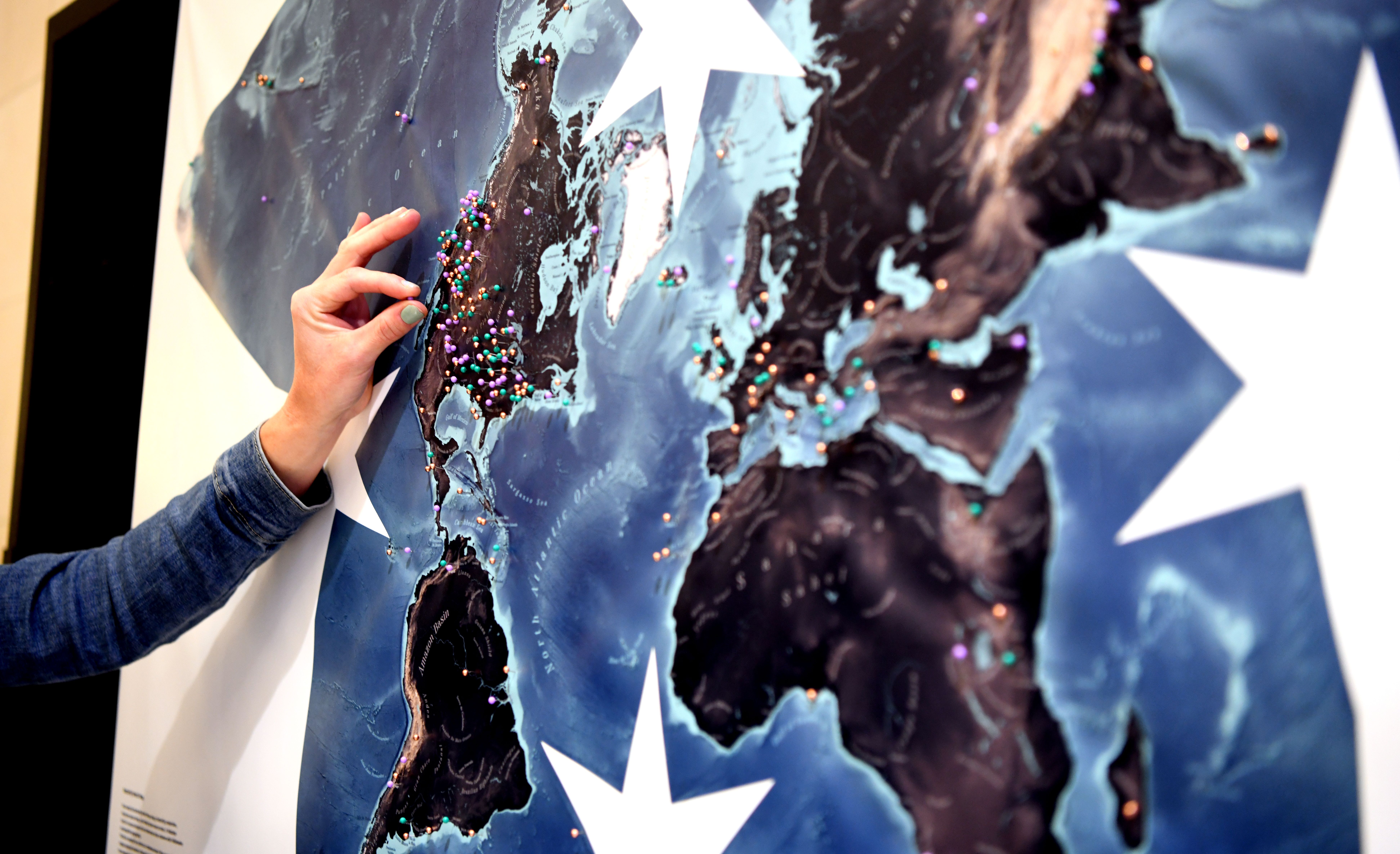 A close-up of a hand placing a colorful pin on a large stylized world map display covered in multicolored pins marking locations across the globe at GSA Connects 2025.