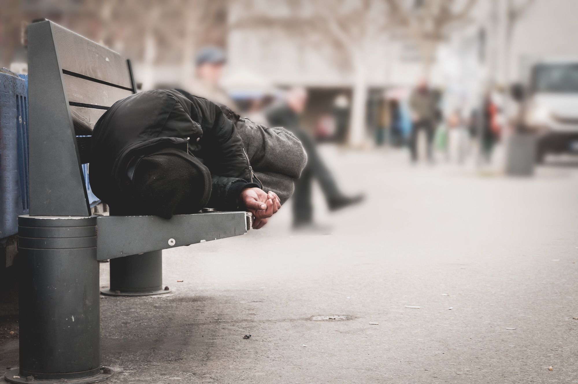 A person sleeping on a public bench as pedestrians pass by on a cold city street.